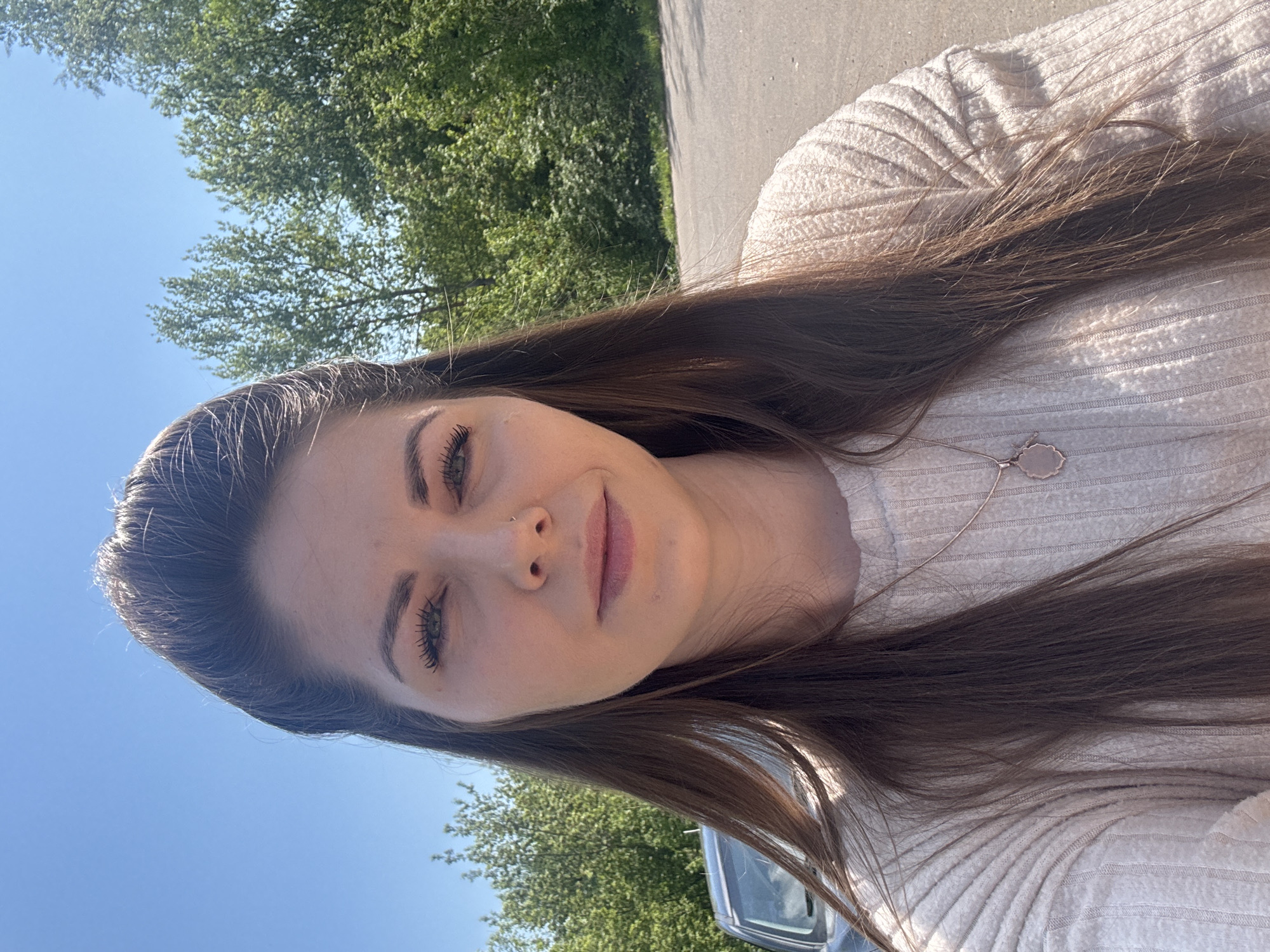 A young woman with long brown hair is posing for a selfie outside. She is wearing a light-colored ribbed sweater and a delicate necklace. The background shows trees, a blue sky, and a glimpse of a car. There is no definitive information in the image to place the location specifically in Shevlyakovo, Russia.