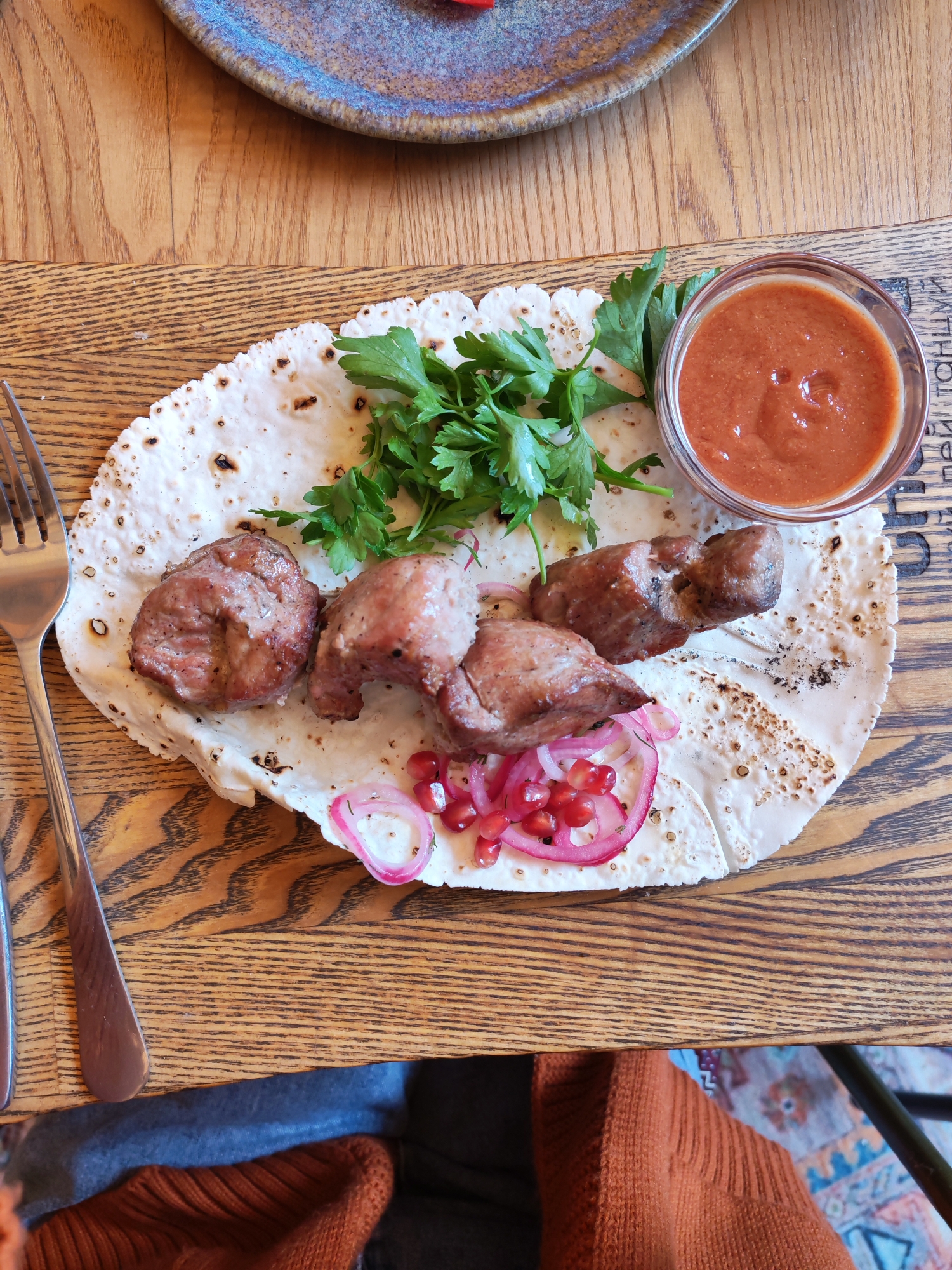 A close-up, overhead shot captures a serving of grilled meat skewers with lavash bread, garnished with parsley, thinly sliced red onion, and pomegranate seeds, served on a wooden board. A small glass bowl of reddish-orange sauce sits to the right of the food. A fork is visible on the left side of the frame. The wooden table has a prominent grain pattern. In the background, a portion of a blue and brown patterned rug is visible, along with a glimpse of dark fabric, possibly clothing. The scene is set in Bol'shie Mkhi, Russia.