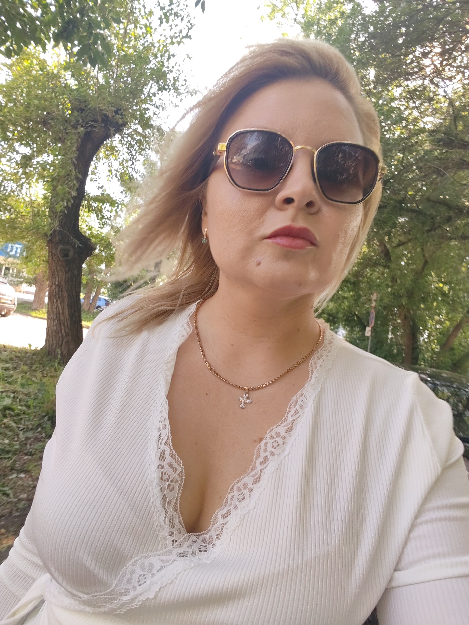The image is a selfie taken outdoors in the city of Izhevsk, Russia. A woman with blonde hair and sunglasses is the main subject. She is wearing a white, ribbed V-neck top with lace trim and a gold chain necklace with a cross pendant. The background consists of trees and foliage, suggesting a park or street with trees. There are cars and buildings visible in the distance. The lighting indicates it is daytime.