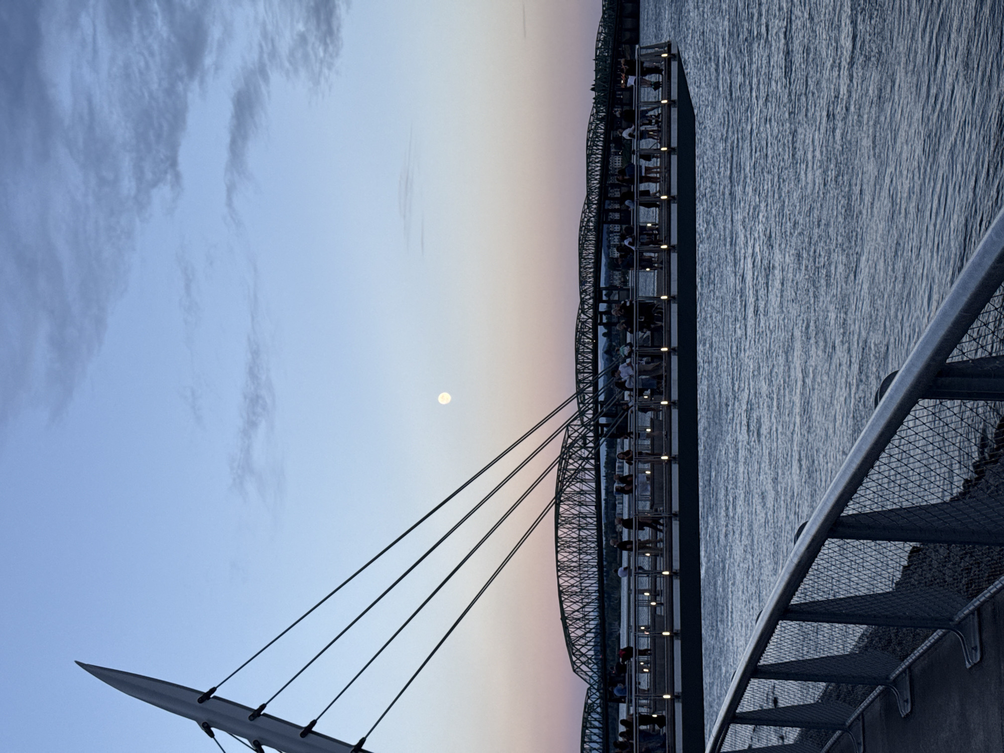 The image depicts the Vancouver waterfront at dusk, likely near the Gastown or Coal Harbour area. A modern pedestrian bridge, characterized by its distinctive mast and suspension cables, spans across the foreground. Several people are visible on this bridge, observing the twilight sky and the water. In the background, a long, multi-arched bridge crosses the water, with more structures and city lights suggesting the urban landscape of Vancouver. The sky transitions from a soft blue at the top to a warm pink and orange hue near the horizon, indicating sunset or sunrise. The moon is visible as a pale orb in the upper portion of the sky. The water is calm, reflecting the muted colors of the sky and the lights from the bridges and the pedestrian walkway. A metallic railing with a wire mesh infill borders a pathway in the extreme foreground, leading the viewer's eye towards the main scene.