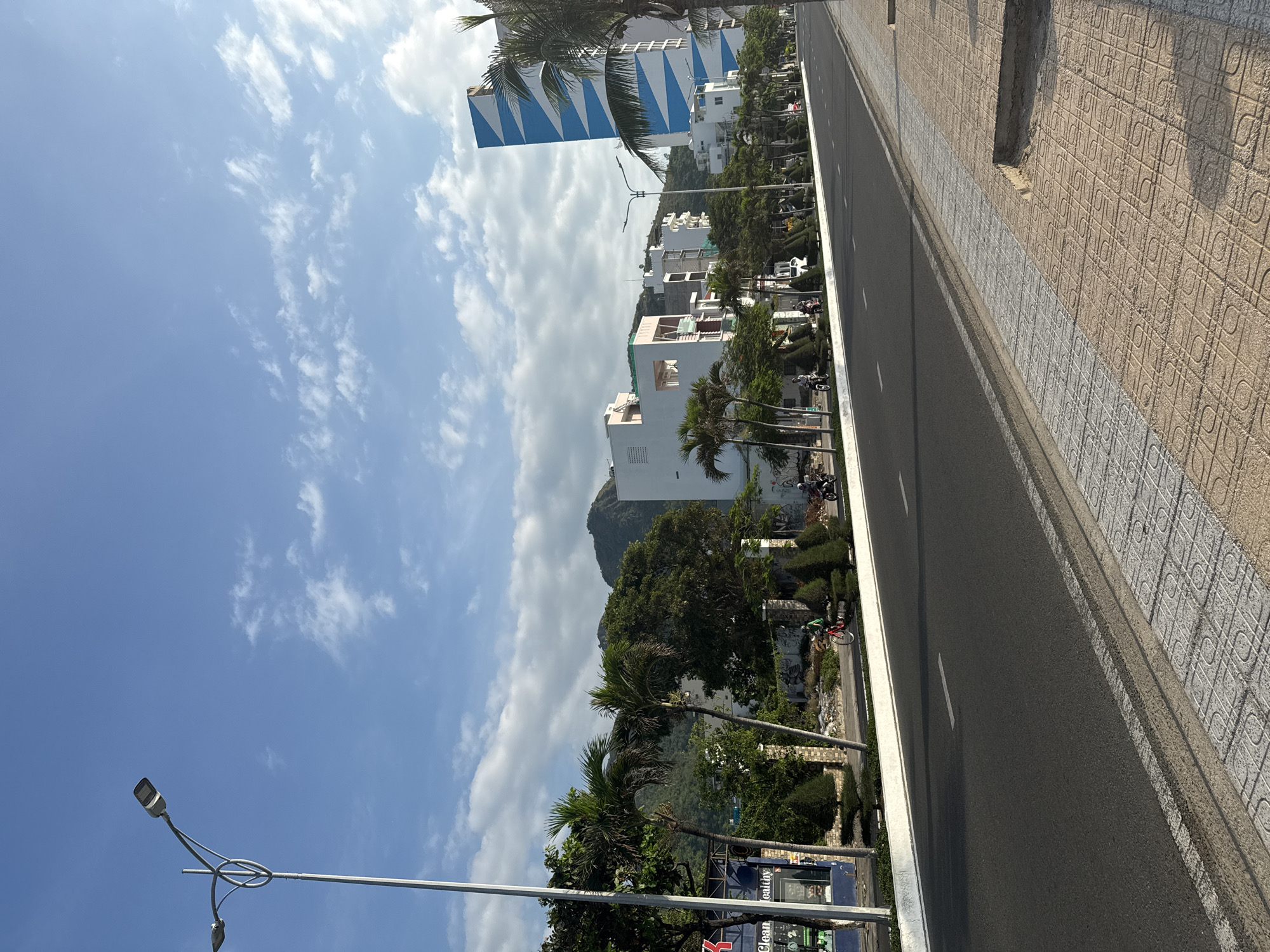 The image depicts a street scene in Bac Nha Trang Ward, Vietnam, on a clear day with scattered clouds. A wide, paved road runs through the center of the frame, with a sidewalk to the right. The sidewalk is paved with square tiles in a light brown and gray pattern. The road is dark asphalt, with white lane markings.
On the left side of the road, a sidewalk area is lined with manicured green bushes and trees, including several palm trees. Behind the greenery, there are buildings with white facades and some with red accents. A tall street lamp with two lights stands on the left edge of the road. Further down the street, a modern building with a distinctive blue and white striped facade is visible. In the distance, a mountain range is visible, covered in trees.
Several people and vehicles are present. A person on a bicycle, wearing a green shirt, is riding on the left sidewalk. Several motorcycles are also seen on the road, with riders visible on some. The general atmosphere is one of a coastal urban environment with lush vegetation and residential or commercial buildings.