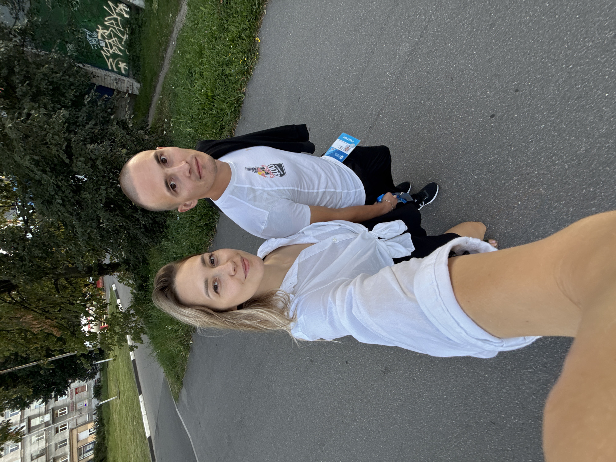 A full-body, slightly angled selfie is taken outdoors. A woman with blonde hair is on the left, smiling and looking at the camera. She is wearing a white collared shirt tied at the waist, black shorts, and black shoes. A man with short, dark hair is on the right, also looking at the camera. He is wearing a white t-shirt with a logo that reads "RED BULL 400 CHAMPIONSHIP," black shorts, and black shoes. He has a black jacket draped over his shoulders and a blue lanyard with a participant card around his neck. They are standing on an asphalt path next to a grassy area. In the background, there is a building with multiple windows, trees, and graffiti on a wall. The scene appears to be in an urban environment, potentially in Saint Petersburg, Russia, given the context of a "Red Bull 400 Championship" which has been held in various locations, including Russia.