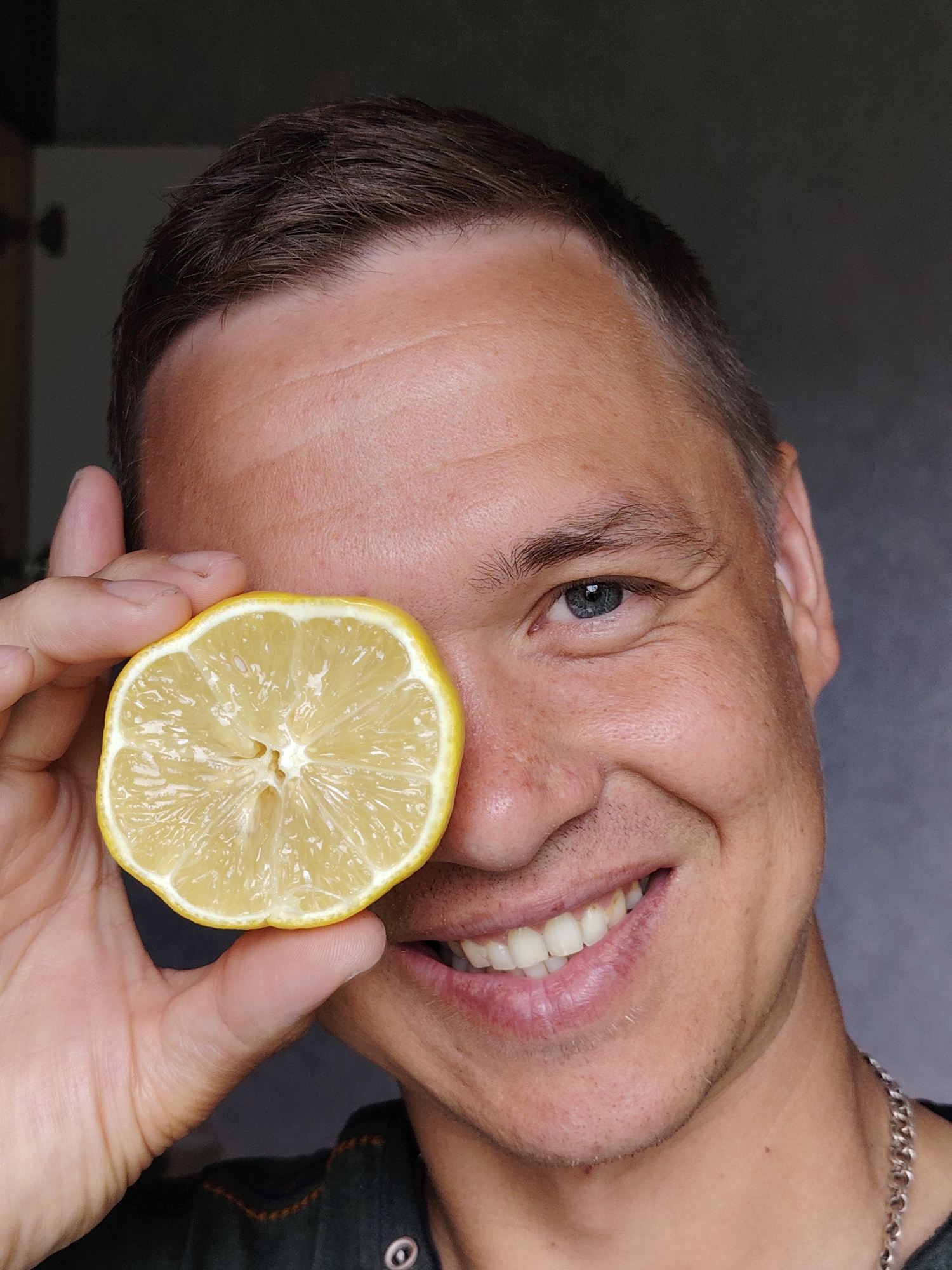 A man is holding a halved lemon in front of his left eye. The lemon is bright yellow with a visible pattern of pulp and seeds. He is smiling, revealing his teeth. The man has short, light brown hair and blue eyes. He is wearing a dark green shirt and a silver chain is visible around his neck. The background is blurred and out of focus, with glimpses of a cabinet or wall. There is no definitive indication of the location being Chaikovsky, Russia in the image.