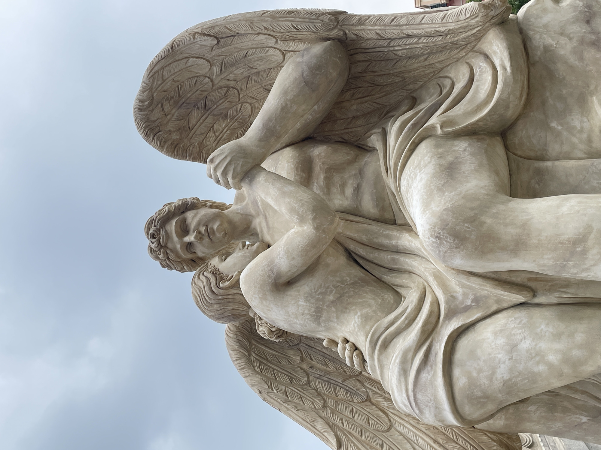 The image depicts a stone sculpture of two winged figures embracing. The figures are rendered in a classical style, with draped clothing and detailed musculature. The larger figure, presumably male, has a muscular torso and is seated, with its arms wrapped around the smaller figure. The smaller figure, leaning into the larger one, has its head resting on the larger figure's chest and its arms around its neck. Both figures have wings, intricately carved to resemble feathers, which extend upwards and outwards. The sculpture is a light, creamy beige color and appears to be made of stone. The background is a pale, overcast sky. This sculpture is located in Nghĩa Trụ Commune, Vietnam, specifically in front of a temple, likely the Bát Tràng Temple.