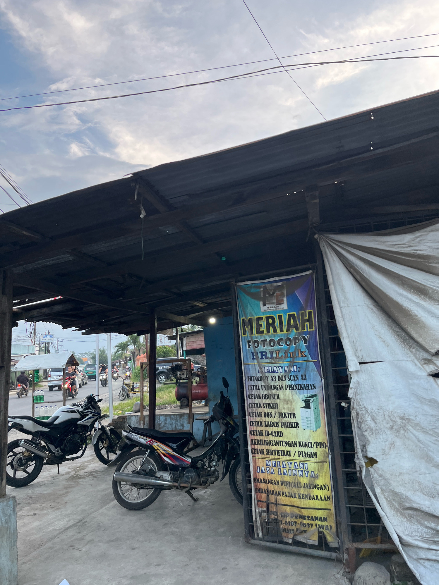 The image shows a storefront in Patumbak Dua, Indonesia. The storefront is covered by a corrugated metal roof and has a tarp on the right side. A sign with the text "MERIAH FOTOCOPY BRILINK" lists services offered, including photocopying, scanning, printing invitations, brochures, stickers, bills, parking tickets, ID cards, keychains, and certificates. There are two motorcycles parked in front of the store, and a few people are visible in the background on motorcycles and in vehicles on a street. The sky is cloudy.