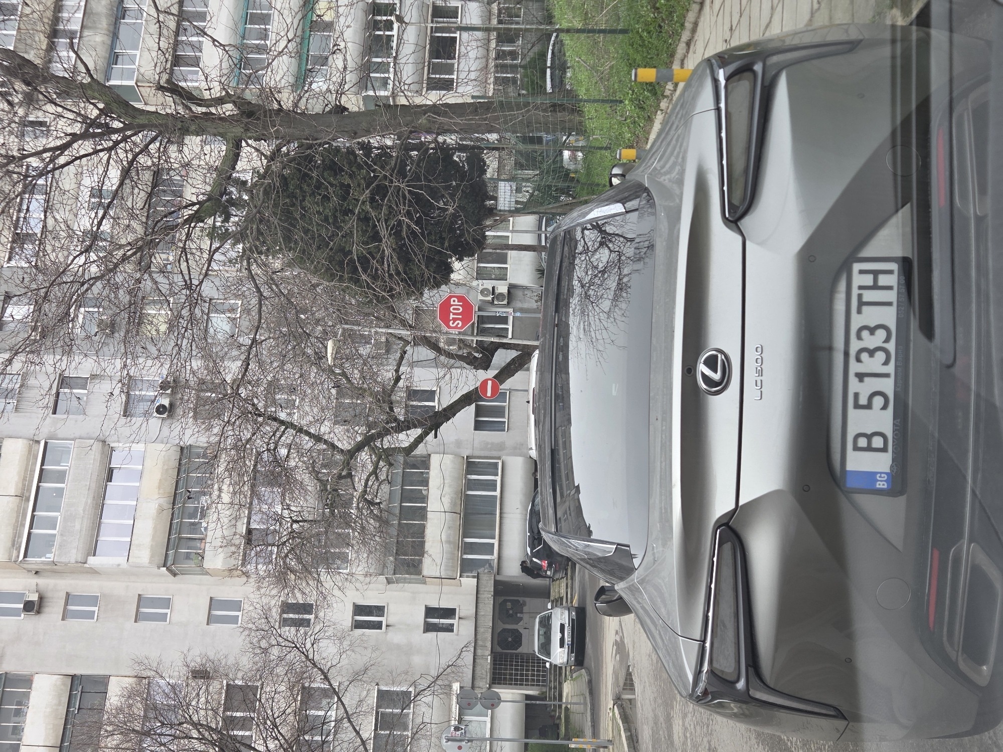 The image depicts a silver Lexus LC 500 with Bulgarian license plate "B 5133 TH" parked on a street in Varna, Bulgaria. The rear of the car is prominently featured. A red stop sign is visible in the background, partially obscured by bare tree branches. The background consists of a large, weathered, multi-story apartment building. A white Skoda car is parked further down the street. The environment suggests an urban residential area, likely during late winter or early spring given the lack of foliage on the trees.