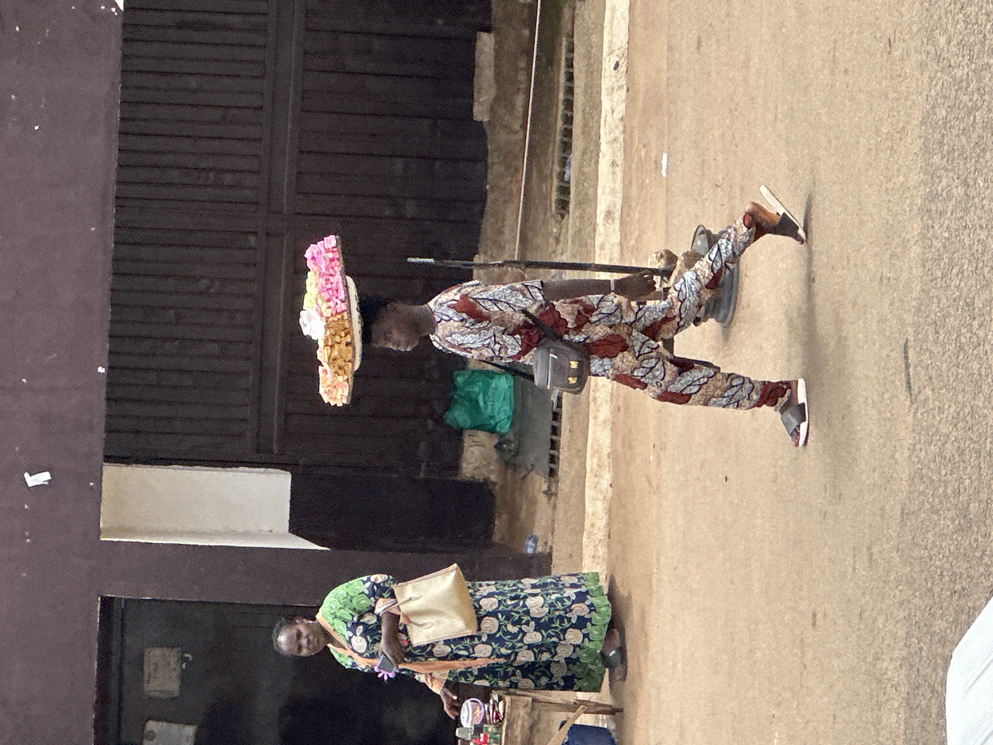 In Bukuru, Nigeria, a young man walks with a tray of colorful sweets balanced on his head. He wears a patterned shirt and trousers, with a black crossbody bag slung over his shoulder. To his left, a woman in a long, floral dress stands behind a small table laden with various items, including drinks and what appear to be small packaged goods. She holds a phone in her hand. The scene takes place on a dusty street in front of a building with dark wooden doors and a dark brown wall. A green plastic bag is visible near the wall.