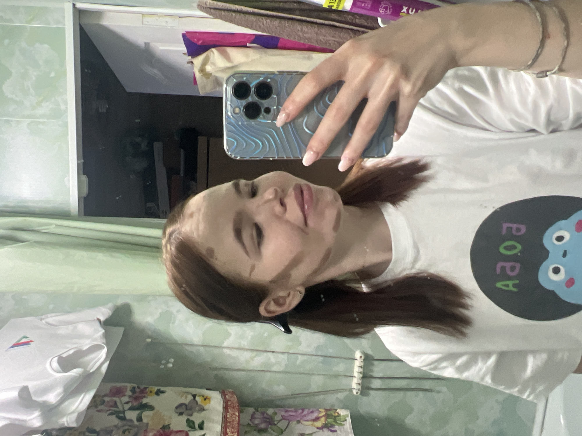 A young woman is taking a selfie in what appears to be a bathroom. She has brown hair with contouring makeup on her face and is wearing a white t-shirt with a cartoon image. The phone she is using has a distinctive patterned case. The bathroom has green walls, a mirror reflecting a cluttered shelf, and other items typical of a domestic bathroom setting, including a clothesline with clothespins. The photo was taken in the city of Chaikovsky, Russia.