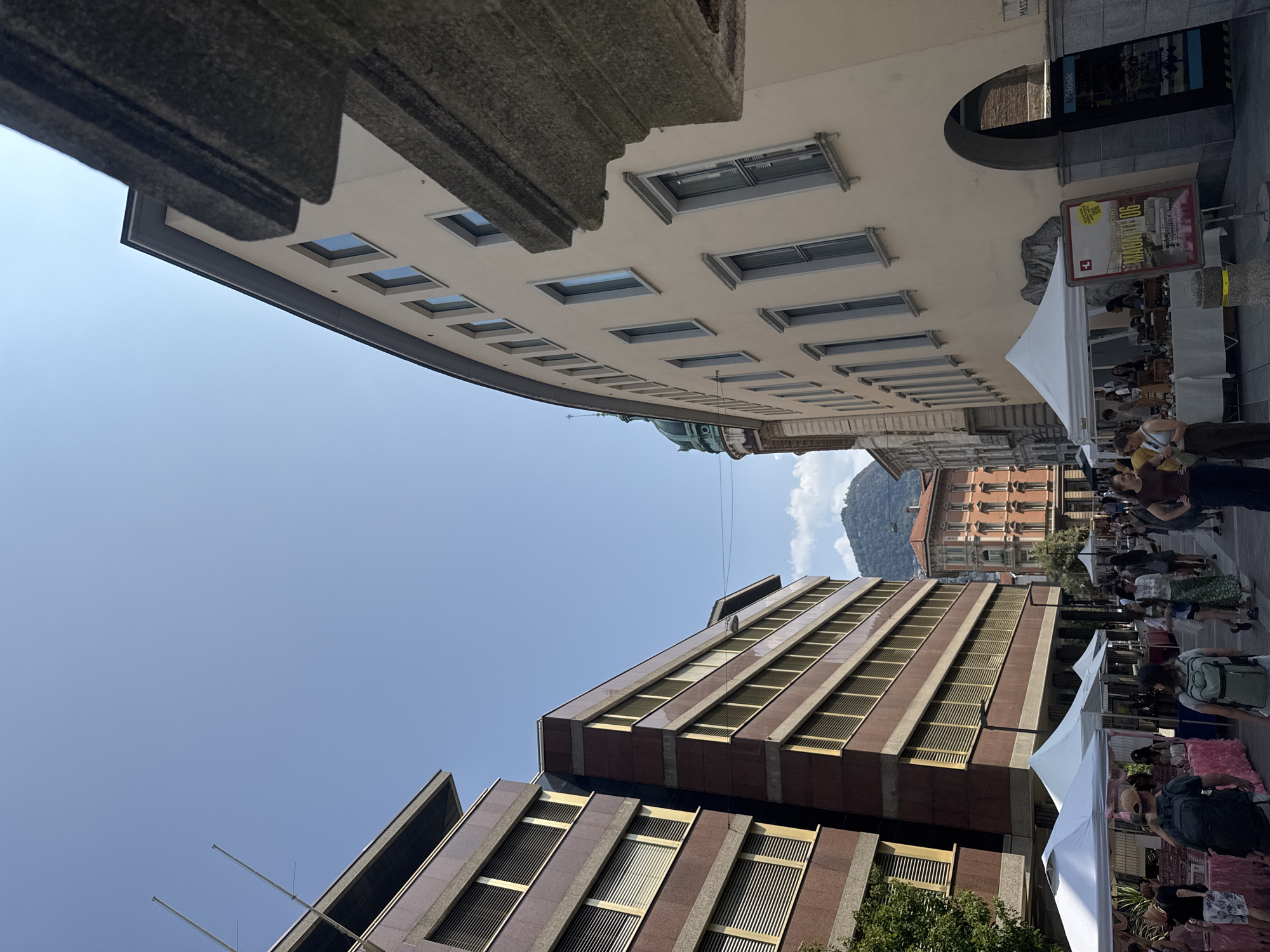 In Lugano, Switzerland, a street market is taking place on a sunny day under a clear blue sky. The scene is dominated by modern buildings with dark red granite facades and beige stucco exteriors, featuring rows of windows with blinds. In the foreground, white tents are set up, displaying goods for sale and attracting shoppers. People are browsing the market stalls, walking along the street, and interacting with each other. In the background, a verdant mountain rises majestically against the sky, adding a natural element to the urban landscape. The architecture suggests a contemporary European city, and the presence of market tents indicates a vibrant public space. The overall impression is one of a lively and pleasant day in the city.