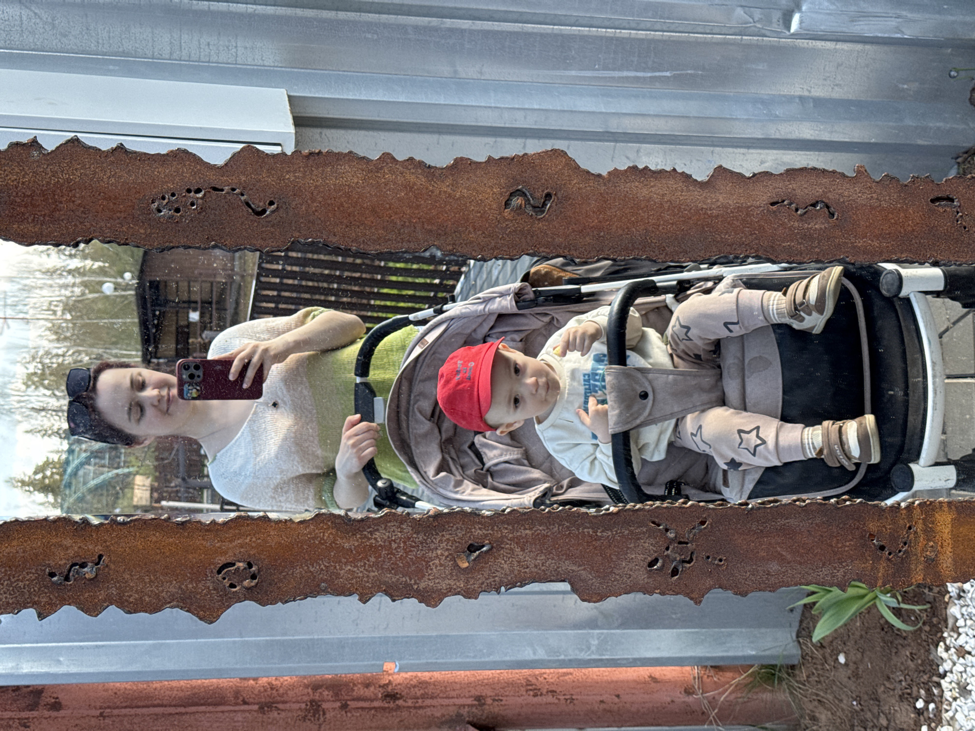 A mirror shot depicts a woman and a baby in a stroller. The woman is holding a phone, presumably taking the photo. The baby is wearing a red cap and pointing. They are positioned in front of a metallic structure with rusted metal trim. The specific location is not identifiable from the image.