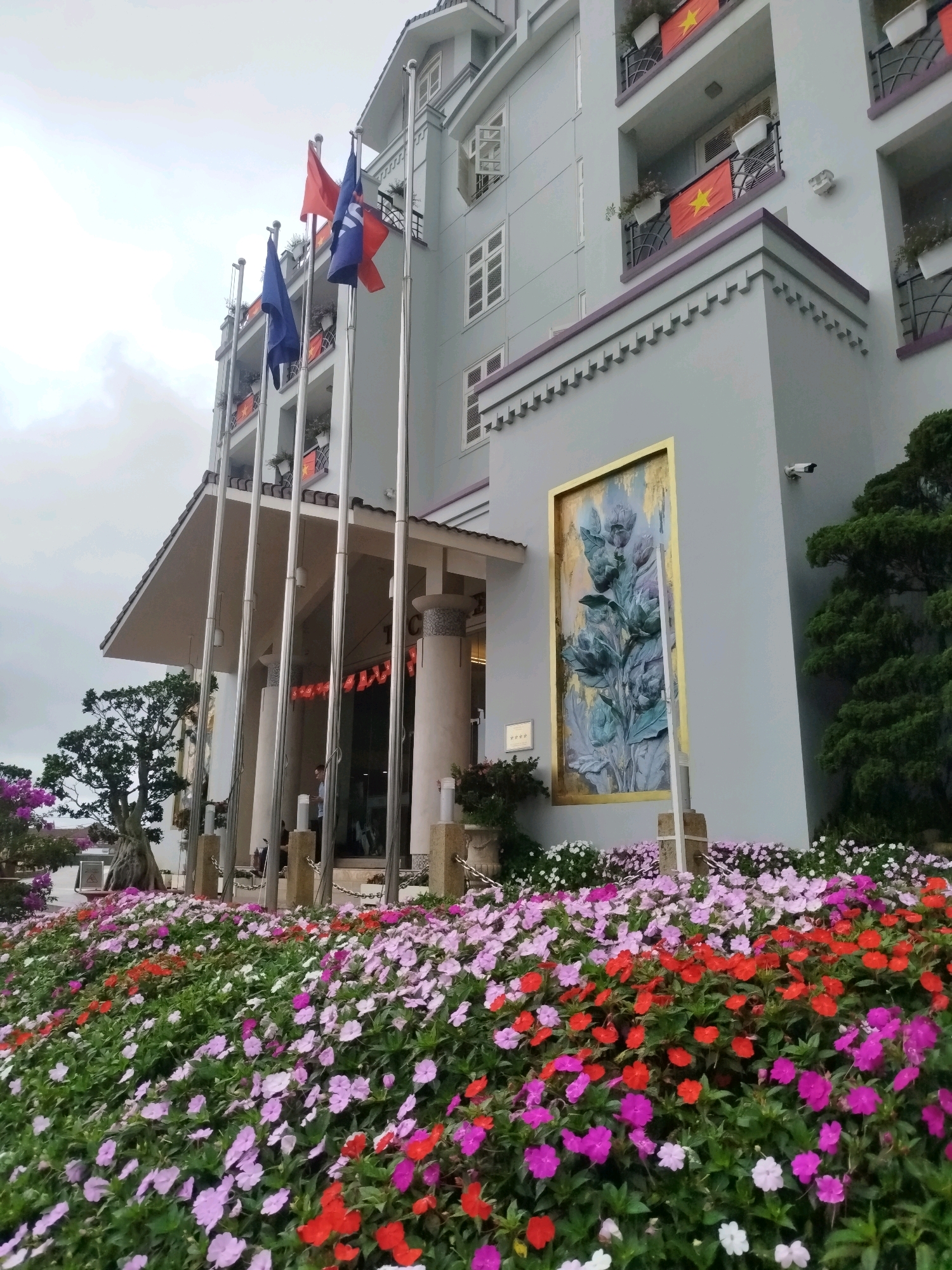 This image captures the exterior of a building, likely a hotel, in Da Lat, Vietnam. The building is light gray with multiple stories, featuring balconies adorned with Vietnamese flags. In front of the building, a vibrant flower bed showcases a profusion of pink, purple, and red impatiens flowers. Several flagpoles stand in front of the entrance, some displaying flags, though their insignia are not entirely clear. A large, framed artwork with a floral motif is mounted on the building's facade. The scene is set against an overcast sky. The presence of the Vietnamese flags strongly suggests a patriotic theme or location.