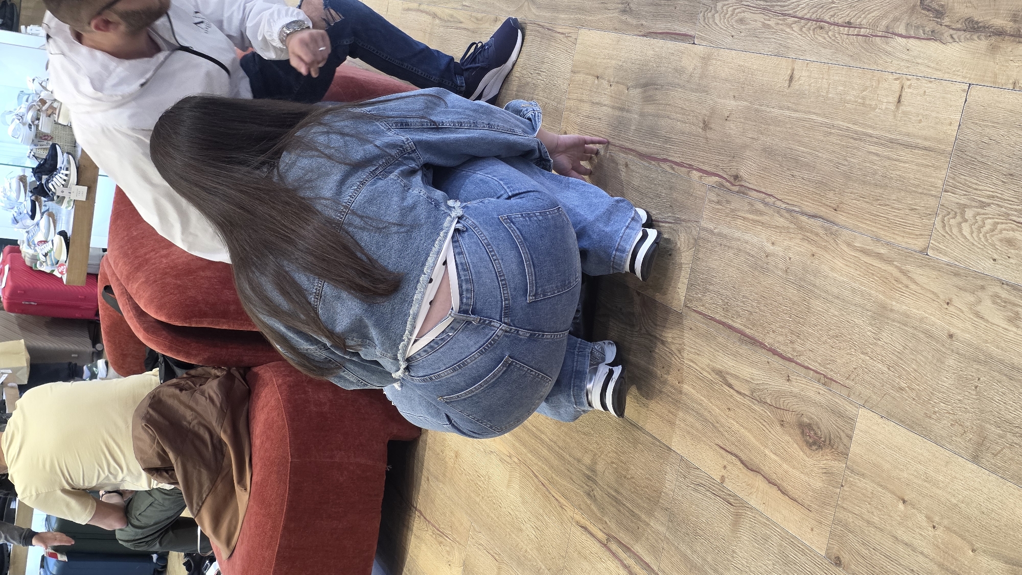 A view from above shows several people seated on a red sofa, with wooden flooring in the background.
One person, dressed in a white jacket and blue jeans, is partially visible. Another person is seen wearing a denim jacket and matching jeans with white and black striped socks. Part of someone wearing a light-colored shirt and brown jacket is also in the frame.
The scene appears to be indoors. Beyond the sofa, shoes and suitcases are visible, which implies the location may be related to travel, perhaps a waiting area in the city of Blagoevgrad, Bulgaria.