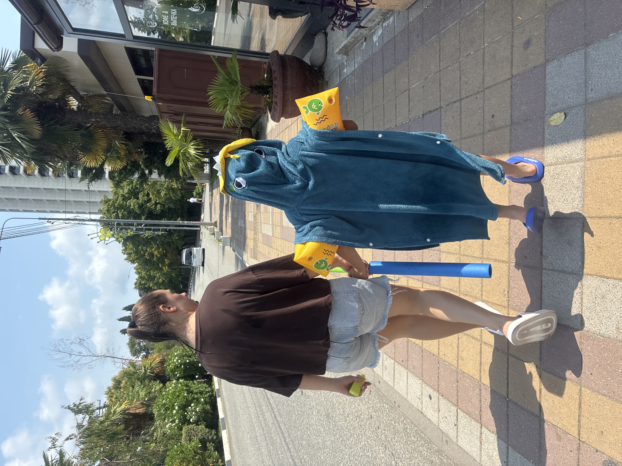 Two people walk along a paved sidewalk in Sochi, Russia, on a sunny day. The person in front, a child, wears a blue towel that resembles a dinosaur costume with a yellow hood and yellow arm floats. The child holds a blue foam pool noodle. The person behind, an adult woman, wears a brown t-shirt and denim shorts. She holds a yellow object in her left hand. The sidewalk is paved with square tiles in shades of brown, tan, and beige. A road runs alongside the sidewalk, with a white van visible in the distance. Lush green trees and palm trees line the street, and a tall, modern building is visible in the background. A building with large windows, possibly a cafe, is on the right side of the frame, with a sign that appears to read "КОФЕ & ВЫПЕЧКА" (Coffee & Pastries).