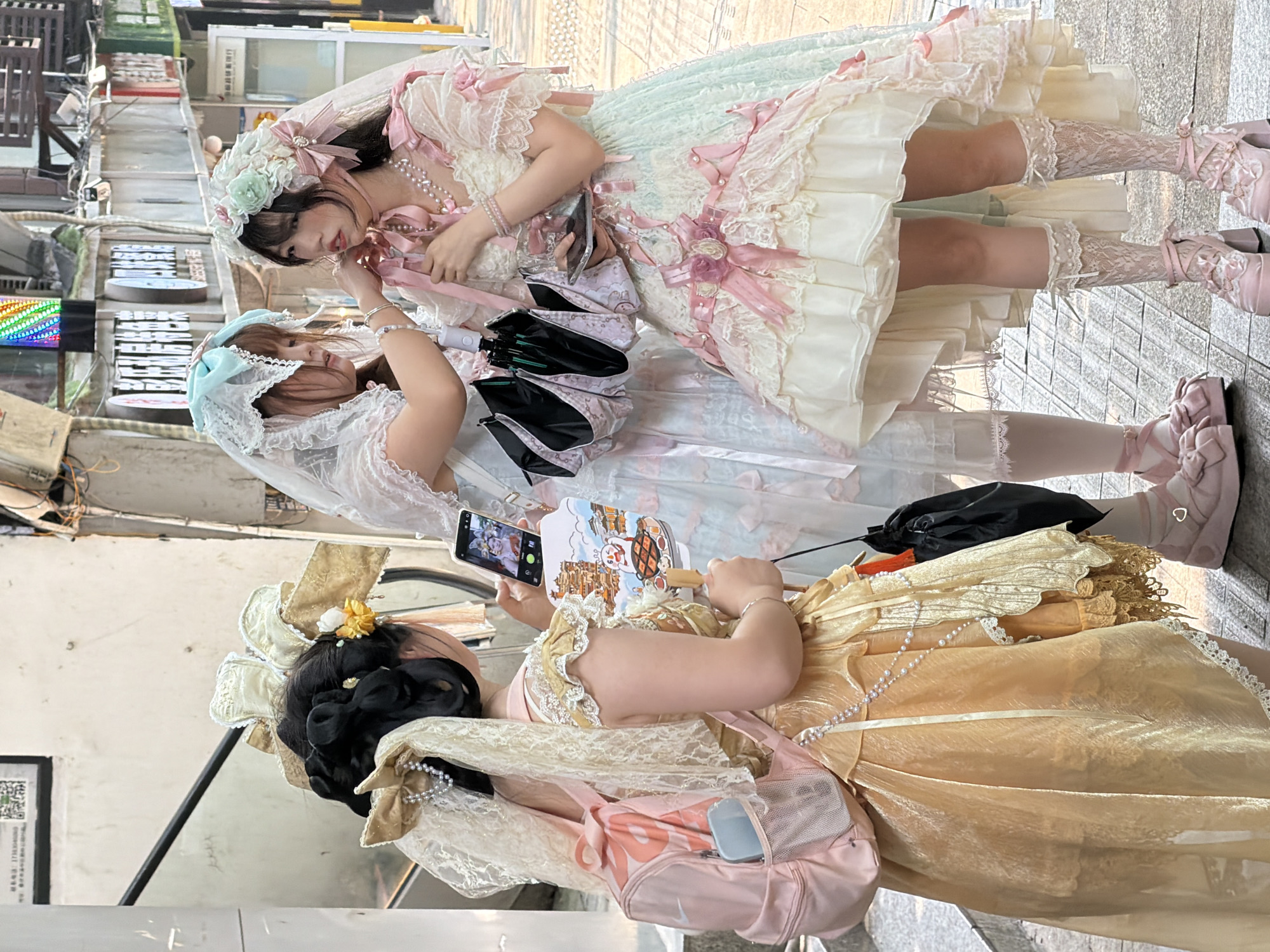 Three young women, dressed in elaborate Lolita fashion, are observed standing on an urban street in Yuzhong District, China.
The leftmost individual, viewed from the back, wears an elaborate golden-hued, multi-tiered dress with lace and ruffled detailing. Her black hair is styled in an ornate bun adorned with large golden bows, lace, and strands of pearls. She carries a pink Nike mini-backpack with a light blue smartphone visible in its mesh side pocket. In her right hand, she holds a white hand fan with a wooden handle, illustrated with a cartoon image of a building and a character. Her left hand holds a smartphone, displaying a screen image of two individuals, presumably a photograph.
The central figure wears a white and light blue tiered dress featuring lace details and pink ribbons. She is veiled with light blue lace and a matching large bow atop her head. A silver bracelet is present on her left wrist. She holds a closed, patterned pink umbrella with a black interior. Both her hands are engaged in adjusting an accessory near the face of the rightmost individual. She wears light pink platform shoes with bows and light-colored lace socks.
The rightmost individual is attired in a multi-layered dress with a white bodice, extensive pink ribbon embellishments, and a mint green and white ruffled skirt with lace overlays. She wears multiple strands of pearl necklaces and a pink bracelet on her left wrist. Her head is adorned with an elaborate headpiece consisting of a white veil, green and pink floral elements, and pink bows. She holds a small, transparent rectangular object in her right hand. Her footwear consists of elaborate pink velvet-like platform shoes with multiple straps and bows, paired with white lace socks.
The background features a light-colored building wall with a diagonal black handrail, suggestive of an escalator or staircase. High on the left wall, a black sign displays a QR code and Chinese characters. Further to the right, indistinct shop signs with Chinese characters and a vertical, multicolored LED light fixture are visible. The ground is paved with grey, rectangular interlocking stones.