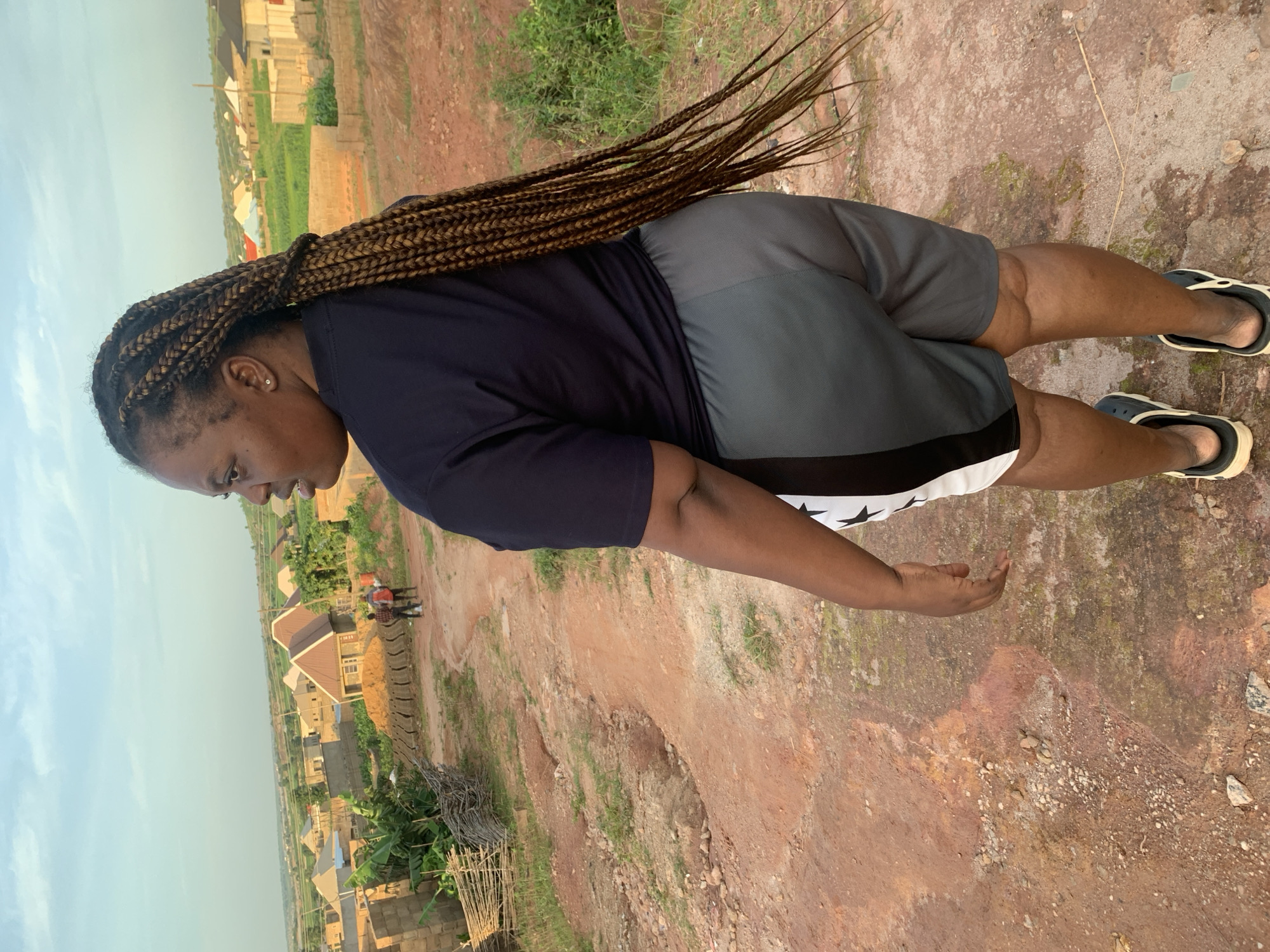 A medium shot captures a Black woman from the posterior as she looks left, standing on a reddish-brown dirt path that slopes upwards. She has long, braided dark brown hair that cascades down her back. She is wearing a dark blue t-shirt and grey shorts with white and black star accents. The background reveals a hazy overview of a village, likely Rido, Nigeria, characterized by numerous buildings with diverse roof colors, including brown, white, and blue. Green vegetation is interspersed among the structures, and a few people are visible in the distance, appearing as small figures on the path. The sky is a light blue with wispy clouds. The terrain is uneven, with scattered rocks and patches of dry grass.
