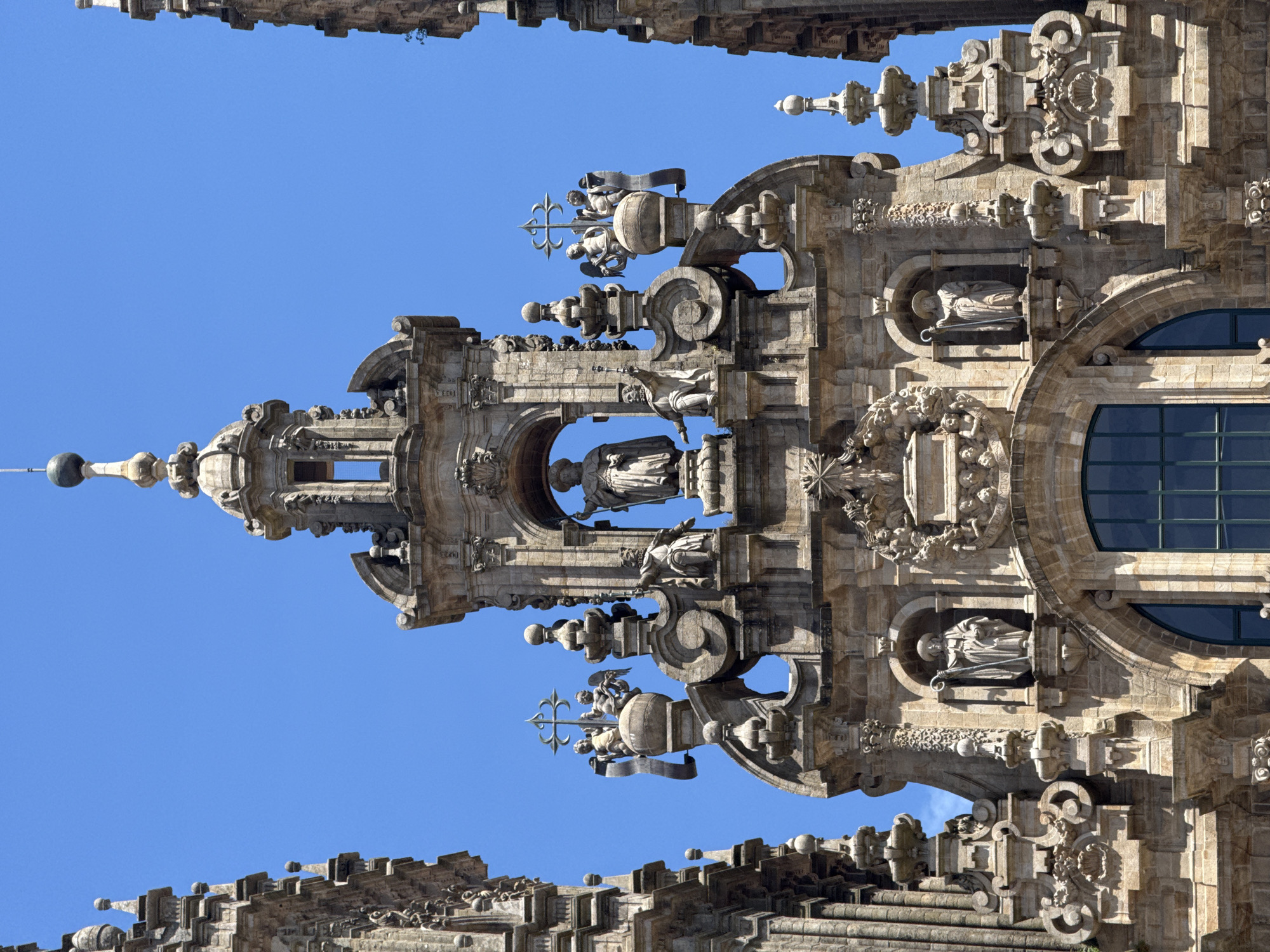 This is a vertical, close-up shot of a detailed stone facade of a building, likely a cathedral given the ornate carvings and statues. The architecture appears to be in a Baroque or Renaissance style, with intricate sculptural elements, arches, and columns. A large, arched window with dark panes is visible on the right side of the frame.
The facade is adorned with multiple statues, including figures in niches and on pedestals. One prominent statue appears to be a seated figure within a rounded niche, while others depict figures in more dynamic poses, some with angelic features. At the top center of the image, a pointed spire-like structure culminates in a cross or fleur-de-lis emblem, flanked by figures. Another section shows what appear to be cherubs or putti holding a decorative banner. The stone itself is weathered, showing the texture and age of the building.
The bright blue sky in the background indicates a clear day, likely during daylight hours, possibly mid-morning or afternoon due to the shadows cast on the intricate stone carvings, highlighting their three-dimensionality. The overall impression is one of grandeur and historical significance. The image is rotated 90 degrees counter-clockwise.
While no specific text is visible, the elaborate ornamentation and religious imagery strongly suggest a significant religious site, consistent with the provided location context of Santiago De Compostela, Spain, a renowned pilgrimage destination.
