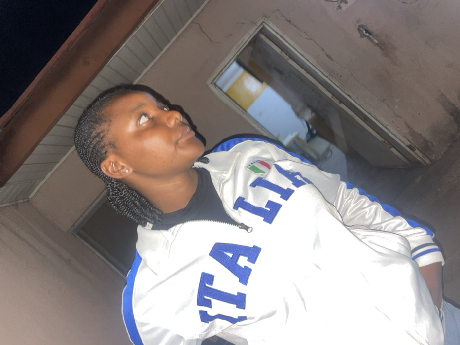 A young woman with dark skin and braids stands indoors, looking upwards. She wears a white jacket with blue accents and the word "ITALIA" prominently displayed. A small Italian flag patch is also visible. The surrounding environment appears to be an unfinished or dilapidated room with exposed beams and a doorway leading to another space. The lighting is dim, casting shadows on the walls.