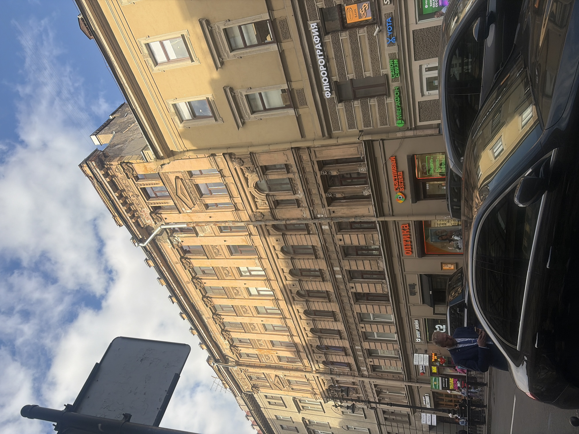 This is a vertical, low-angle shot of a street in Saint Petersburg, Russia. The scene features ornate, old buildings lining the street, their facades weathered and displaying intricate architectural details. Several signs are visible on the buildings, including "ФЛЮОРОГРАФИЯ" (Fluorography), "БАНК" (Bank), "МЕГАФОН" (Megafon), "YOTA", "оптика" (Optics), "счастливый взгляд" (Happy Look), and "ТАБАК" (Tobacco). The sky above is a mix of blue with scattered clouds, suggesting daytime.
In the foreground, a dark car is parked at an angle, reflecting the buildings opposite. A person in a dark suit and blue shirt, possibly a businessman, is standing next to the car, looking at their phone. Another person is visible further down the street, near a sign for "PRICE COFFEE." A traffic sign is partially visible in the bottom left corner, tilted upwards. The overall impression is a glimpse of everyday life on a historic city street, with a blend of modern commerce and traditional architecture. The lighting suggests it is mid-day or late afternoon.