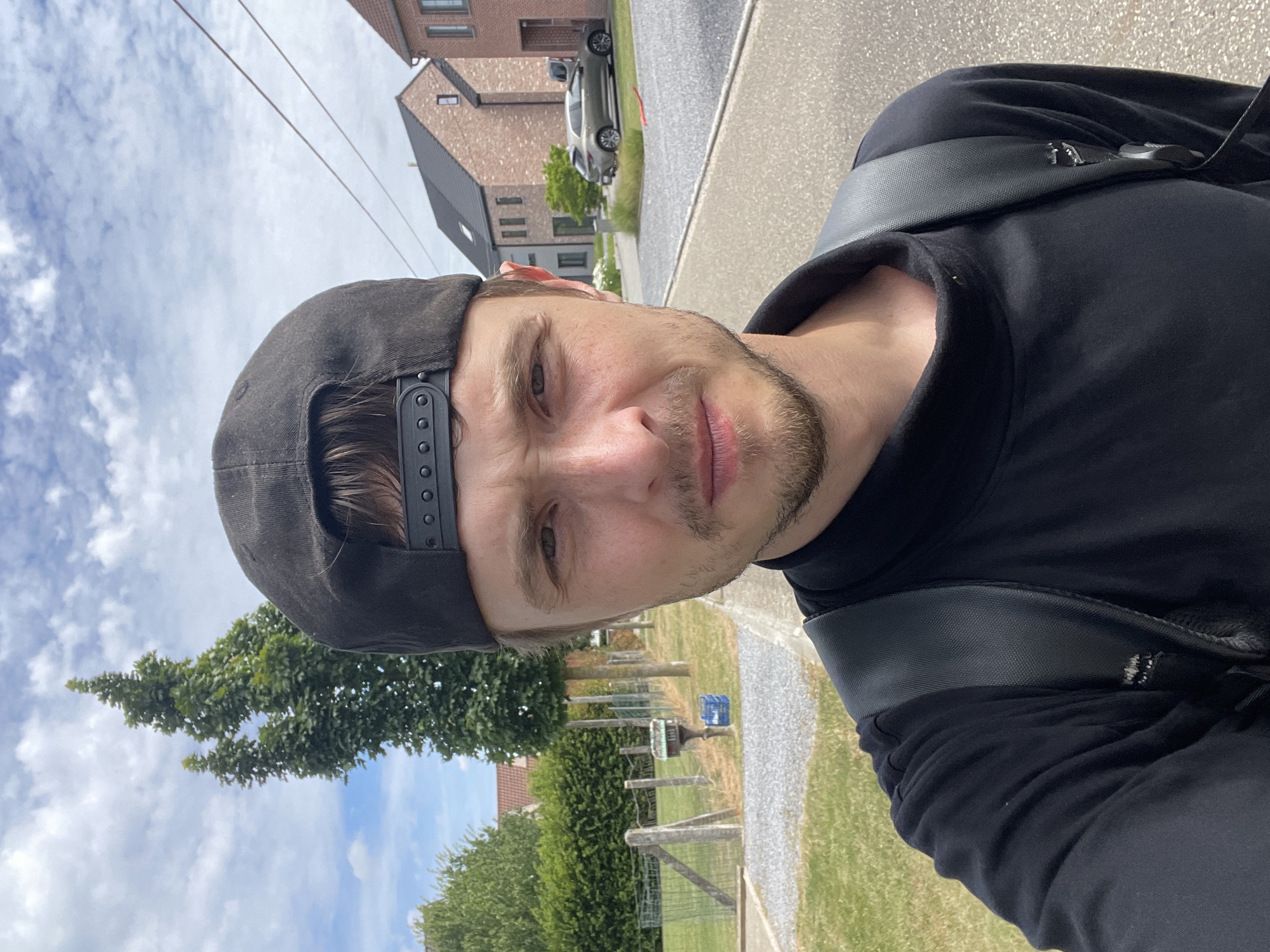 A young man, wearing a black t-shirt and a black baseball cap, stands outdoors, taking a selfie. He has a light beard and stubble. A black backpack is visible on his shoulders. In the background, to the left, a green hedge lines a fence, with a small metal mailbox attached to one of the fence posts. A blue crate sits on the grass next to the mailbox. Further back, there are trees and houses with red-tiled roofs. To the right of the man, a paved road leads past more houses and a parked car. The sky is partly cloudy. The setting appears to be a residential street in Diepenbeek, Belgium.