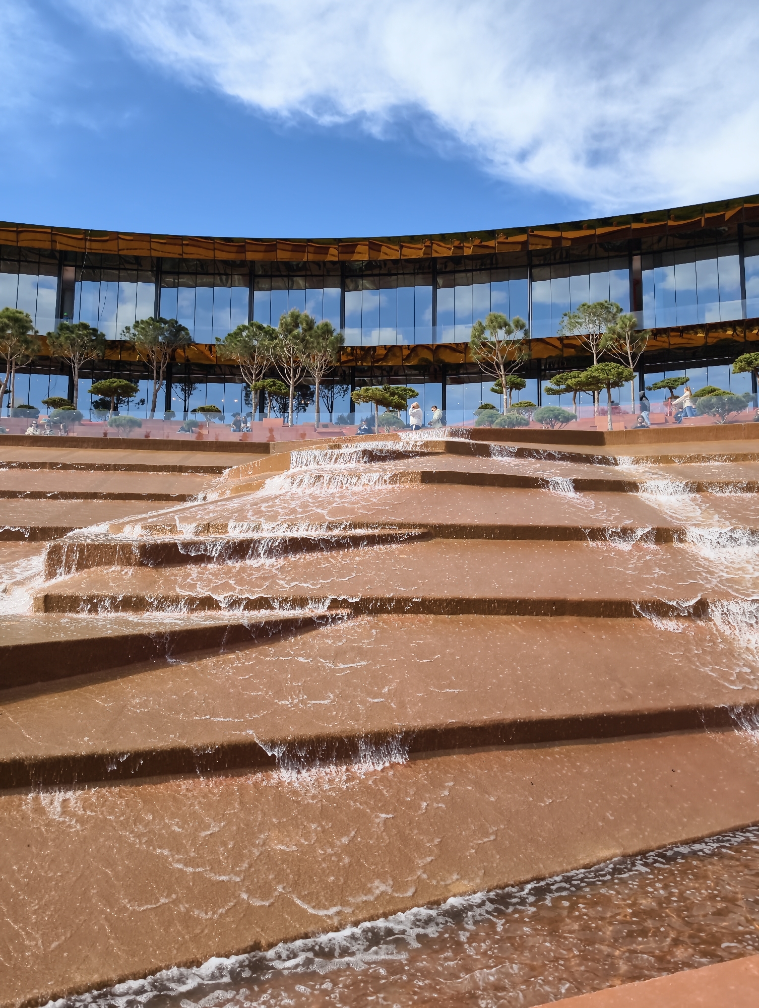 The image displays a modern architectural landscape featuring a large, multi-tiered water feature and a curved glass building, set under a bright blue sky with scattered clouds. The scene is captured during daylight, suggesting fair weather.
In the foreground, a prominent cascade fountain flows down a series of broad, reddish-brown or terracotta-colored steps. Water courses over these steps, creating white foam and ripples, and appears to be slightly brownish, possibly due to sediment or the color of the fountain material. The fountain extends across the lower portion of the frame, drawing the eye upward.
Above the fountain, in the midground and background, a distinctive, contemporary building curves across the horizon. Its facade is primarily composed of large, reflective glass panels that brilliantly mirror the blue sky and white clouds, making the structure blend seamlessly with the environment. The top edge of the building features a unique golden or bronze-toned element, possibly a metal cladding, which adds a warm contrast to the glass and sky.
Along a terrace or walkway in front of the building's glass facade, several small, neatly trimmed trees and shrubs are planted. A handful of people are visible on this terrace, appearing as small figures walking or standing. Their presence suggests the area is a public space, possibly a park or a recreational complex.
The overall impression is one of a meticulously designed urban environment, combining modern architecture with natural elements like water and greenery. There is no visible text in the image. This landscape is characteristic of Krasnodar Park (Galitsky Park) in Krasnodar, Russia.