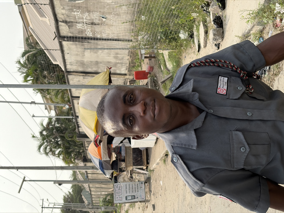 A man in a gray security uniform stands in a sunny outdoor setting. The uniform has a company logo and a decorative rope across the chest. Behind him, there's a street scene with buildings, utility poles, and vendors with umbrellas. The background also features a concrete wall with graffiti. The man is looking towards the camera with a calm expression.
