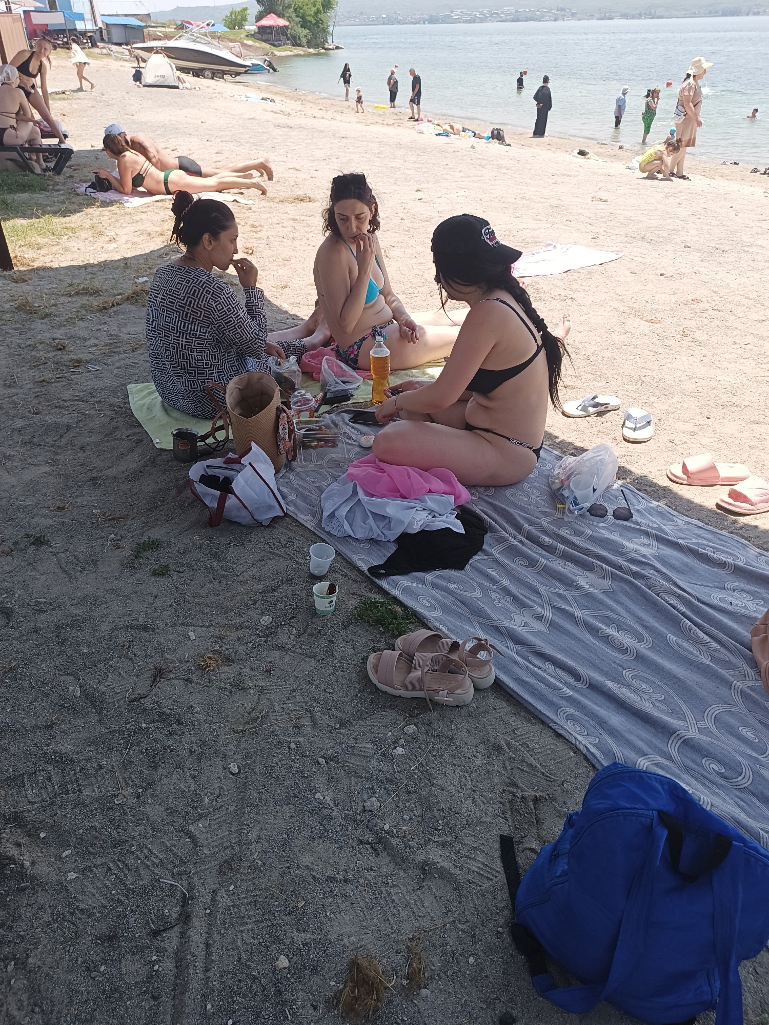 This is a photograph taken on a sunny day at a beach, likely in the city of Sevan, Armenia. In the foreground, three women are sitting on blankets on the sand. The woman on the left is wearing a patterned black and white dress and is holding a food item. The woman in the middle is wearing a blue bikini and is looking down. The woman on the right is wearing a black bikini and a black baseball cap. Various items are scattered around them, including a woven bag, a cooler, plastic cups, a bottle of beverage, and clothing. In the background, other people are visible on the beach and in the water. Some are sunbathing, while others are wading or swimming. A boat is moored near the shore, and a tent is set up on the beach. The environment appears to be a typical lakeside beach with sandy shores and calm water.