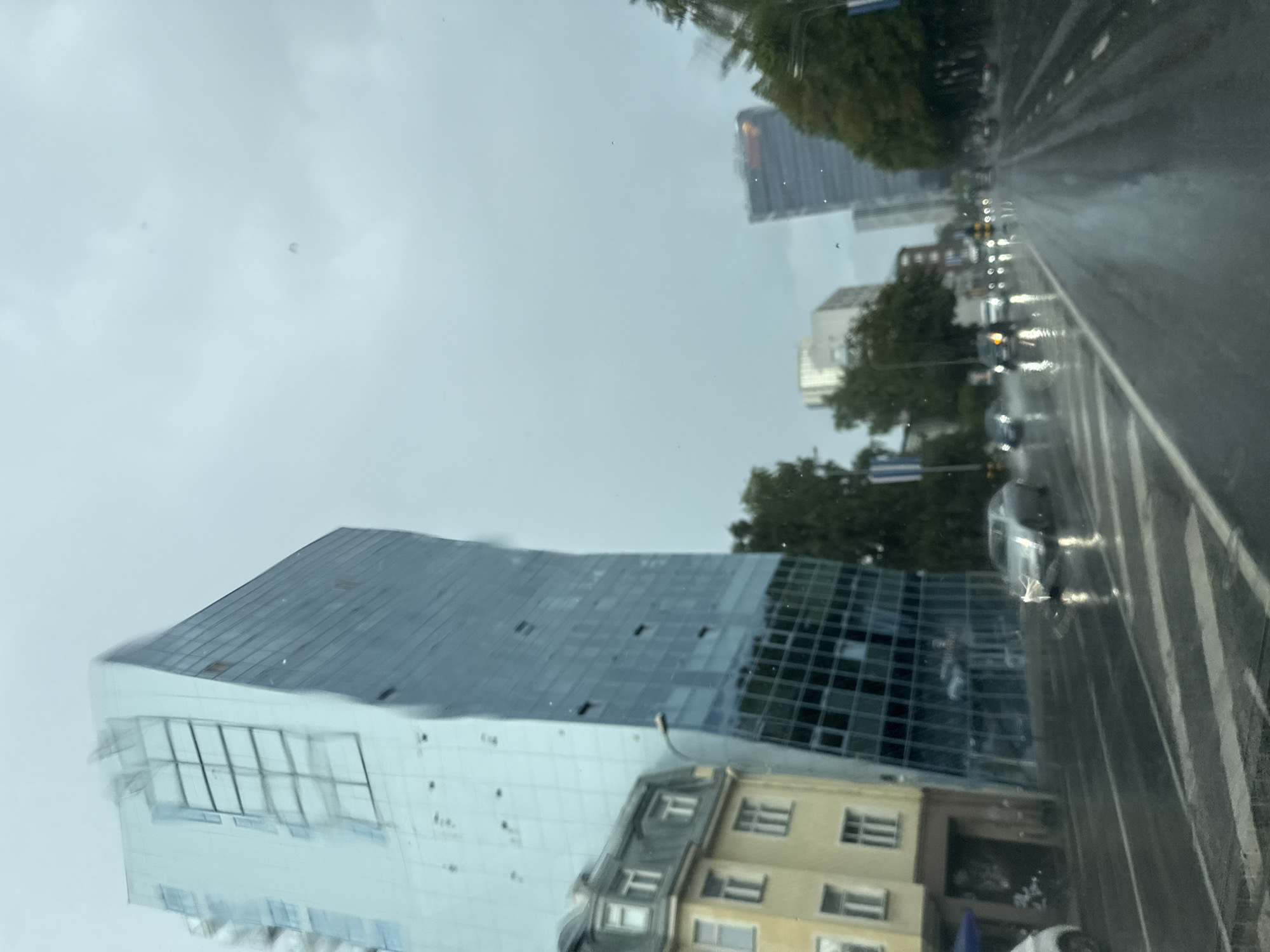 The image depicts a street scene in Tallinn, Estonia, on a cloudy and rainy day. A distinctive, modern building with a curved, glass facade dominates the left side of the frame. To its left, an older, yellow building with a grey mansard roof is visible. The street is wet and reflective from the rain, with several cars driving on it. In the background, other tall buildings and trees can be seen. The overall atmosphere is grey and somewhat subdued due to the weather.