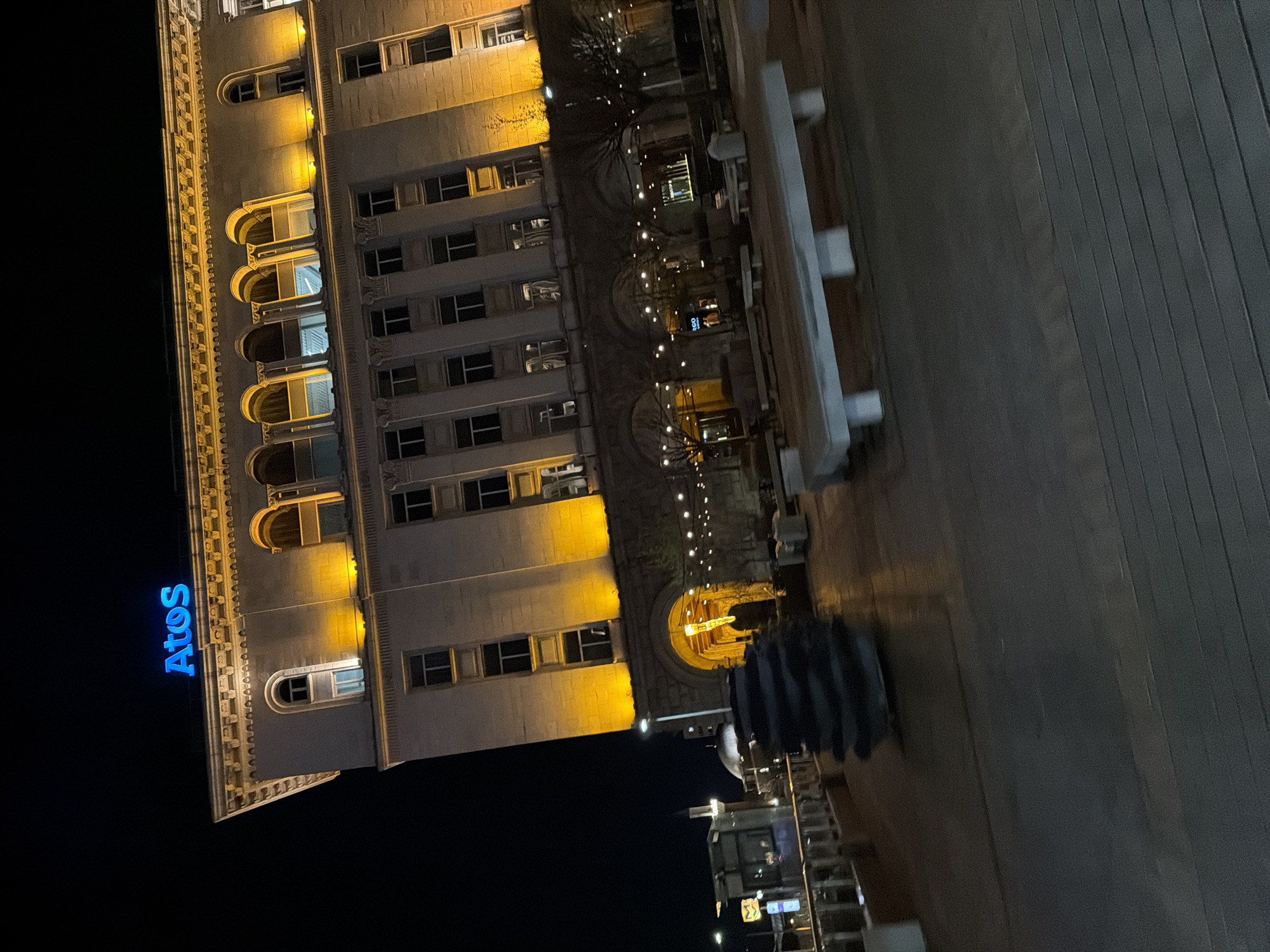 At night, an exterior building with the blue illuminated "Atos" sign is visible on its side, with numerous lit windows and architectural details. Adjacent to the building is a boardwalk. Below is a smaller structure, possibly a food truck. The location is suggested to be Sofia, Bulgaria. No people are clearly visible.