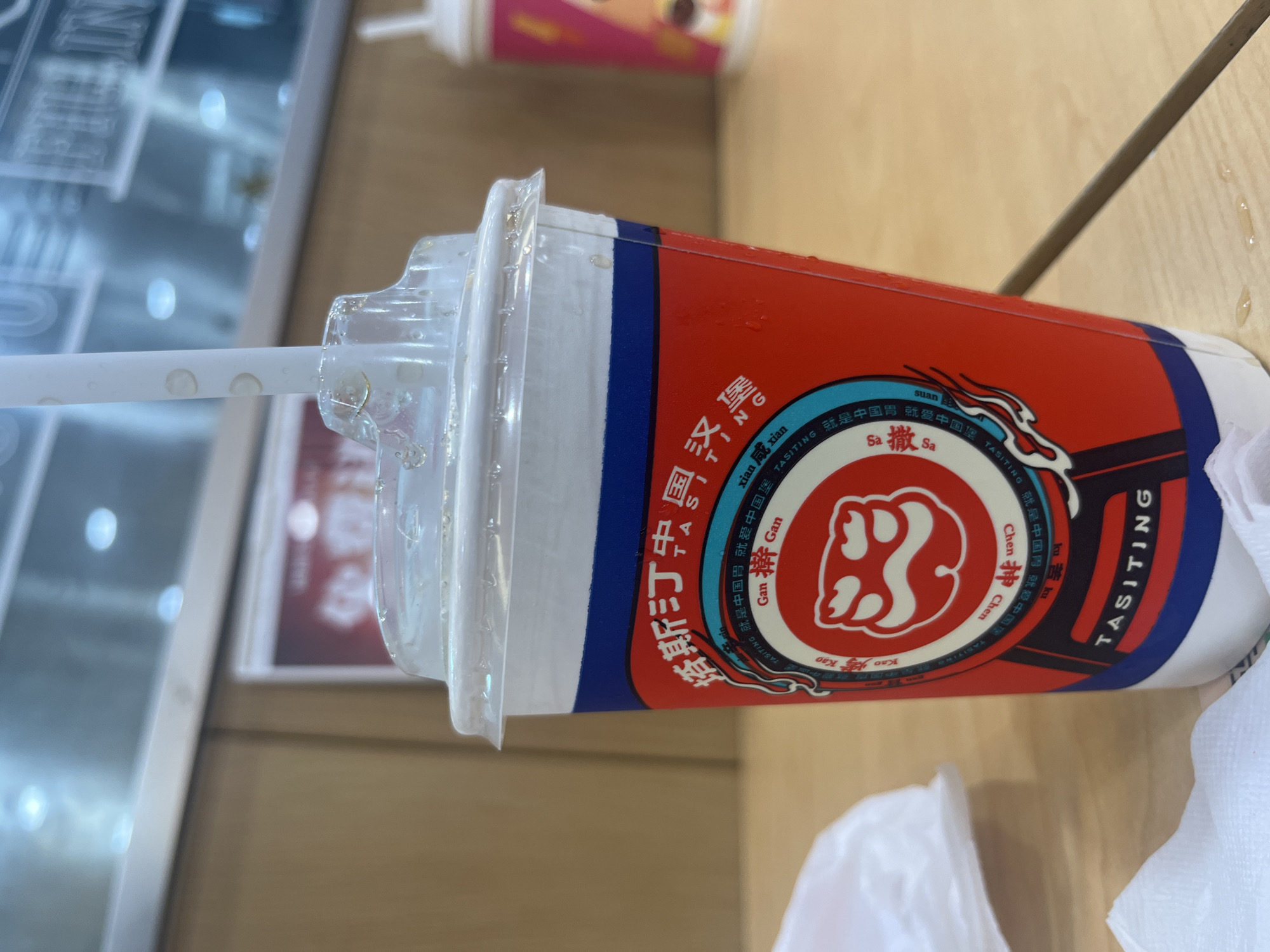 A close-up, eye-level shot shows a red and white plastic cup with a clear plastic lid and straw, sitting on a light-brown wooden table. The cup has a blue band around the top and features Chinese characters and the word "TASITING" printed on it. A circular logo with a stylized white facial expression is visible on the front of the cup. There are water droplets on the lid and cup. To the left, a white plastic bag and a white paper napkin are partially visible. In the background, another blurred cup with a pink and yellow design is present, along with a light-colored wall and ceiling. The image appears to be taken indoors in a commercial setting, possibly a cafe or restaurant, with the overall ambiance suggesting a casual dining environment. The location is Yuzhong District, China.