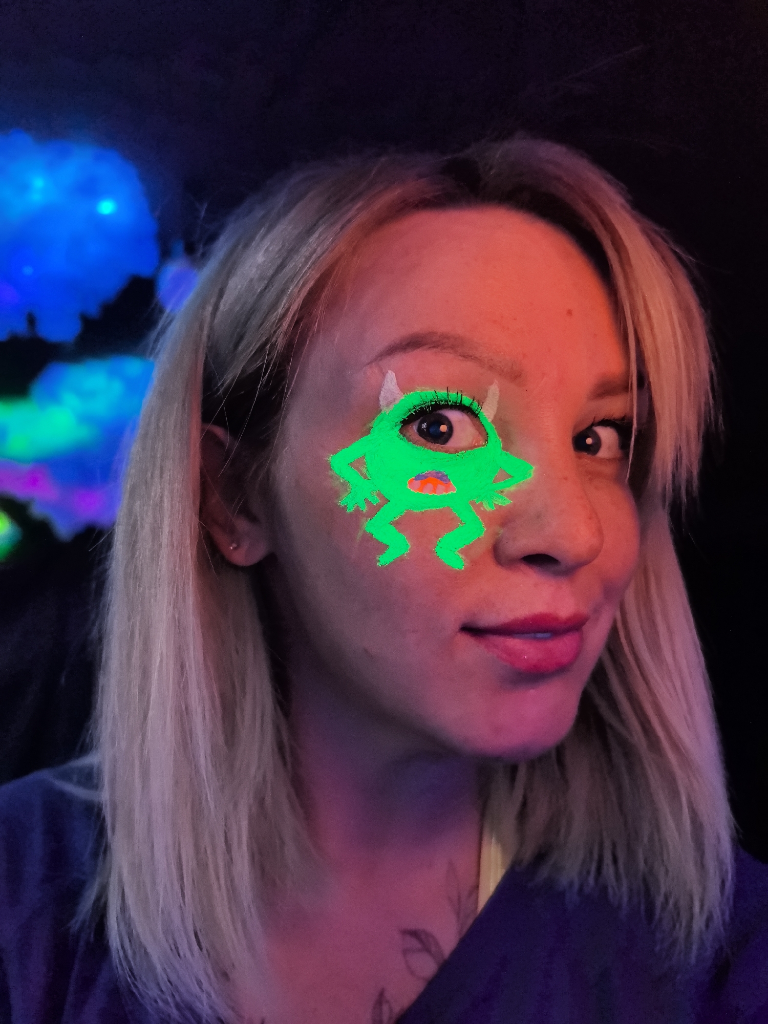 The image captures a close-up portrait of a woman in what appears to be a dimly lit, possibly neon-themed indoor setting in Noviy, Russia. The woman, with blonde hair styled with bangs and pulled back from her face, is looking directly at the viewer with a slight, friendly smile. Her eyes are open and engaging.
A prominent feature is the vibrant, glow-in-the-dark green face paint applied around her left eye (viewer's right). This paint depicts a cartoonish, squat monster-like creature with small horns, arms akimbo, and legs, using her actual eye as the monster's single large eye, complete with drawn eyelashes. A small, red and orange detail is painted on the monster's "body." She is wearing bright pink lipstick and has light makeup around her visible eye.
The lighting in the scene is dramatic and colorful. A strong reddish-orange light illuminates the right side of her face, forehead, and hair, while pink and purple hues are visible on her neck and shoulders. Her blonde hair reflects these various light colors, appearing reddish, pink, and even subtly blue in places. The background is dark, featuring blurry, abstract shapes of glowing blue and green light, reminiscent of cloud-like formations or abstract art installations, suggesting a unique event space, club, or artistic environment. She is wearing a dark, possibly blue or purple, top. There is no visible text in the image.