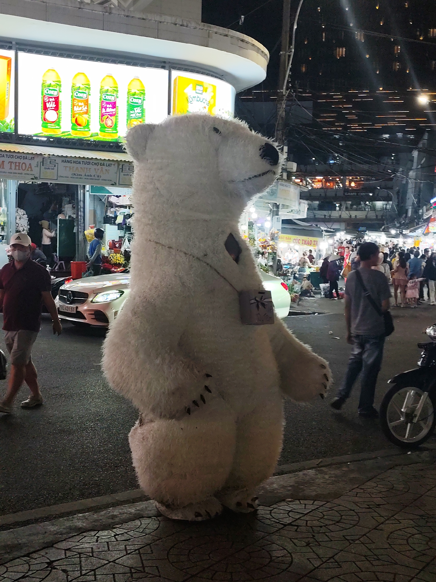 This image depicts a street scene in Da Lat, Vietnam, at night. A person in a large, fluffy white polar bear costume is prominently featured in the foreground, facing to the right of the frame. The costume has a small, gift-like accessory hanging from its chest. To the left of the costume, a man wearing a white cap, dark red polo shirt, and grey shorts walks away from the camera, wearing a face mask. Behind him, a white Mercedes-Benz car is parked, its headlights illuminated. The background reveals a bustling street with various shops and stalls, illuminated by bright lights. Several people are visible walking and standing in the background, and overhead, a network of electrical wires and streetlights is evident. The overall atmosphere suggests a lively urban environment at night, likely a market or commercial area.