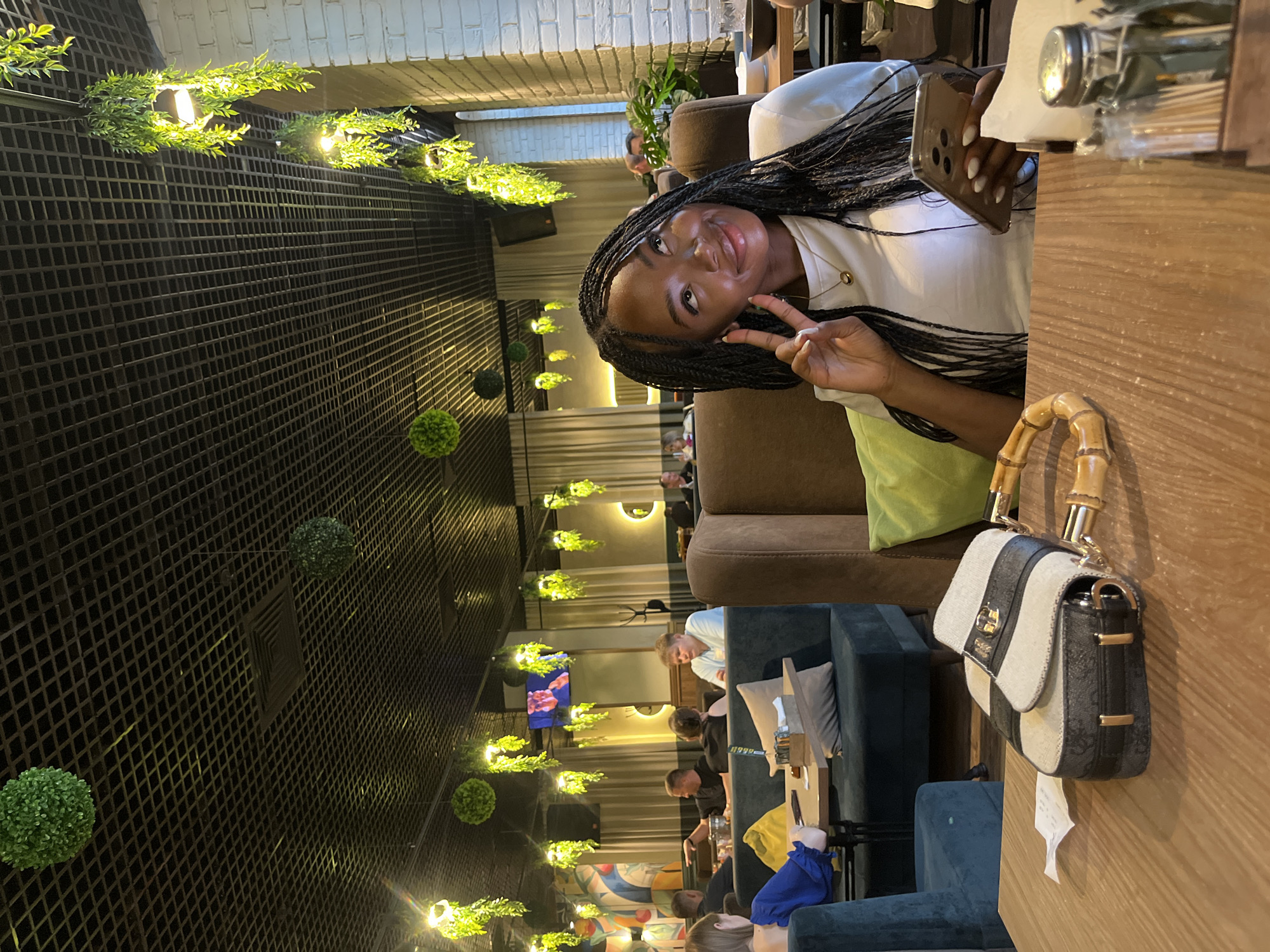 A medium shot captures a young Black woman with long braids seated at a wooden table in a dimly lit restaurant, holding a smartphone. She wears a white t-shirt and a gold necklace. Her left hand is raised near her face, and she appears to be looking upwards. In the foreground, a stylish handbag with bamboo handles rests on the table next to a receipt. The restaurant's interior features a dark, grid-like ceiling adorned with hanging artificial greenery and lights. In the background, other patrons are visible dining at their tables, and a television screen displays an image. The overall ambiance suggests a casual dining establishment.