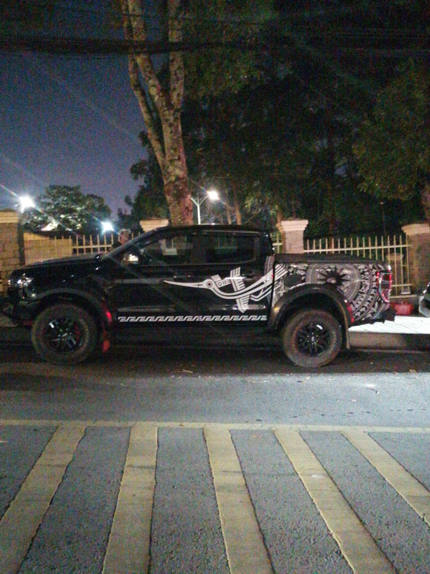 The image shows a black pickup truck parked on the side of a road at night, in Da Lat, Vietnam. The truck is adorned with intricate white tribal-style graphics on its side and bed. The graphics feature geometric patterns and stylized figures, with a particularly elaborate design on the rear quarter panel. The truck has black rims and red brake calipers.
The setting appears to be a street with a pedestrian crossing in the foreground. The road is paved, and the pedestrian crossing is marked with alternating yellow and gray stripes. In the background, there are trees, a fence, and streetlights, suggesting an urban or semi-urban environment. The time of day is clearly night, as indicated by the darkness and the artificial lighting. The weather appears to be clear.
There is no visible activity or event taking place; the truck is stationary. No people are visible in the image. The overall mood is somewhat mysterious and striking due to the contrast of the black truck with the decorative graphics against the dark night. No legible text is present on the vehicle or in the scene.