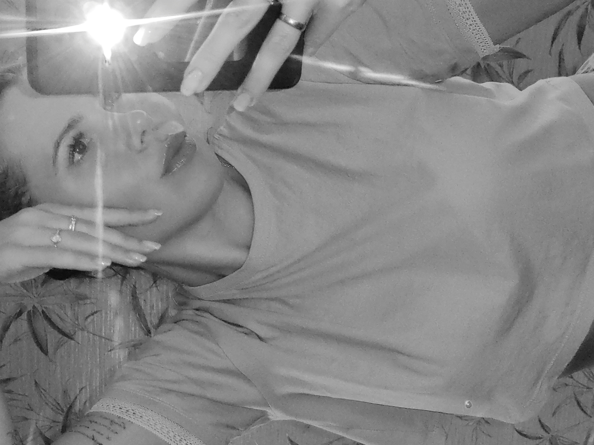 A person lies on a patterned surface, possibly a bed or floor, in a selfie-style photograph. The person holds a phone aloft, its flash creating a glare. They wear a light-colored t-shirt and have rings on their fingers. The photo is black and white and there is no identifying information to determine that it was taken in Chaikovsky, Russia.