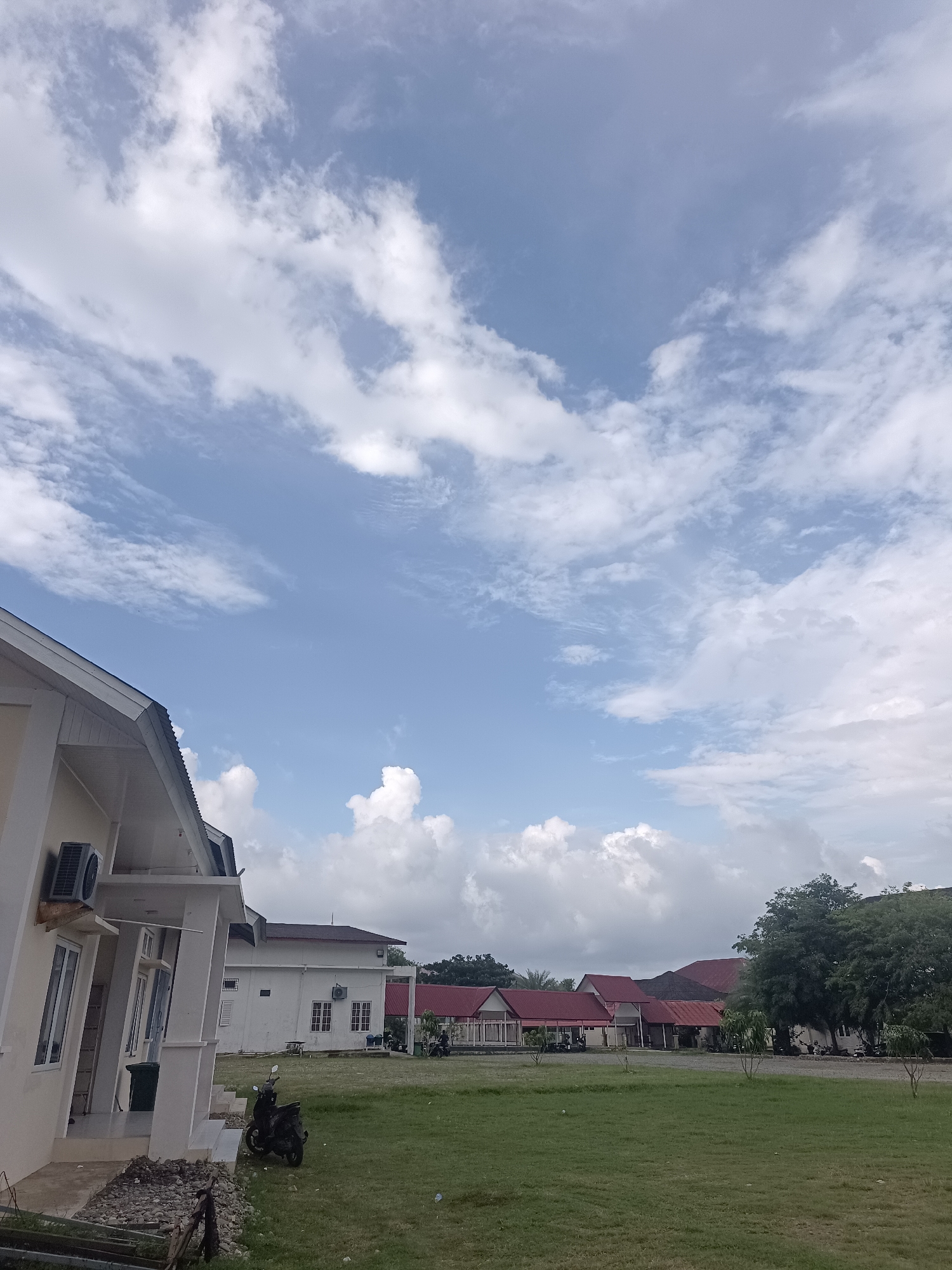 The image depicts a daytime outdoor scene in Mesjid Reuntoh, Indonesia, characterized by a bright blue sky with scattered white clouds. In the foreground, a light-colored building with a white roof is partially visible on the left, featuring an air conditioning unit mounted on its exterior wall. A dark-colored motorcycle is parked on a patch of green grass in front of the building. Further into the scene, a row of buildings with red roofs stretches across the middle ground. These buildings appear to be low-rise structures. Several trees, including a large, mature tree on the right side, are present, and the ground is covered with green grass. The overall impression is that of a tranquil, possibly institutional or residential area under a partly cloudy sky.