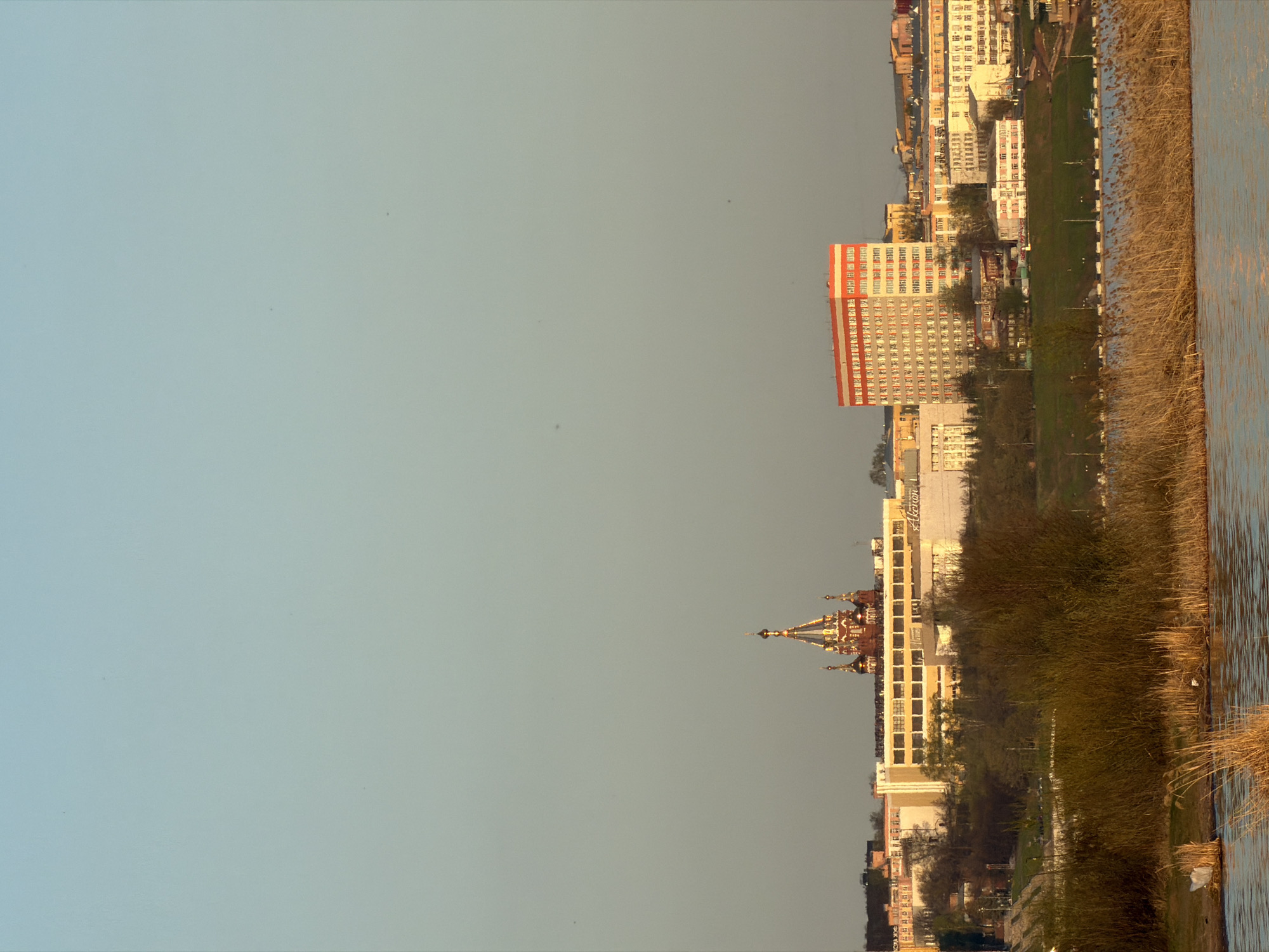 The image shows a cityscape in Izhevsk, Russia, captured from a low-angle perspective. The buildings and infrastructure are vertically oriented on the right side, with the sky on the left. The buildings include a tall, red and white rectangular building, and a distinctive church. The river is to the right. Tall grasses, grow at the water's edge. The sky is clear and of uniform color.