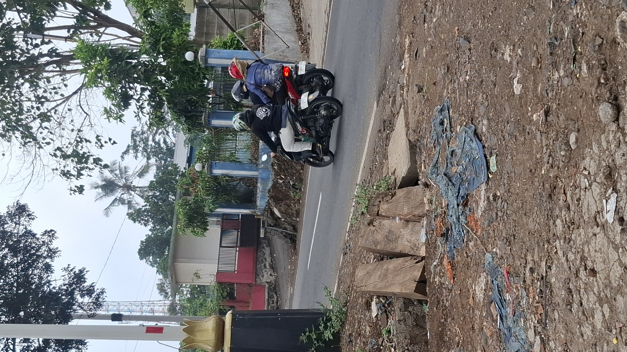 In Cilaku, Indonesia, a side view captures two motorcycles traveling on a paved road under an overcast sky. The lead motorcycle, a black scooter, carries a person wearing a black hoodie and white pants, their face obscured by a helmet with green and white patterns. Behind them, a red scooter is ridden by a person wearing a blue jacket and red helmet, with a second person also wearing a red helmet seated behind them. The road passes by a blue and white fence with decorative pillars, beyond which lies a building and dense foliage. The foreground of the image shows a dirt and gravel embankment littered with debris, including blue tarps, plastic, and pieces of wood. The overall scene depicts a typical Indonesian street environment, marked by both the presence of transportation and the common issue of roadside litter.