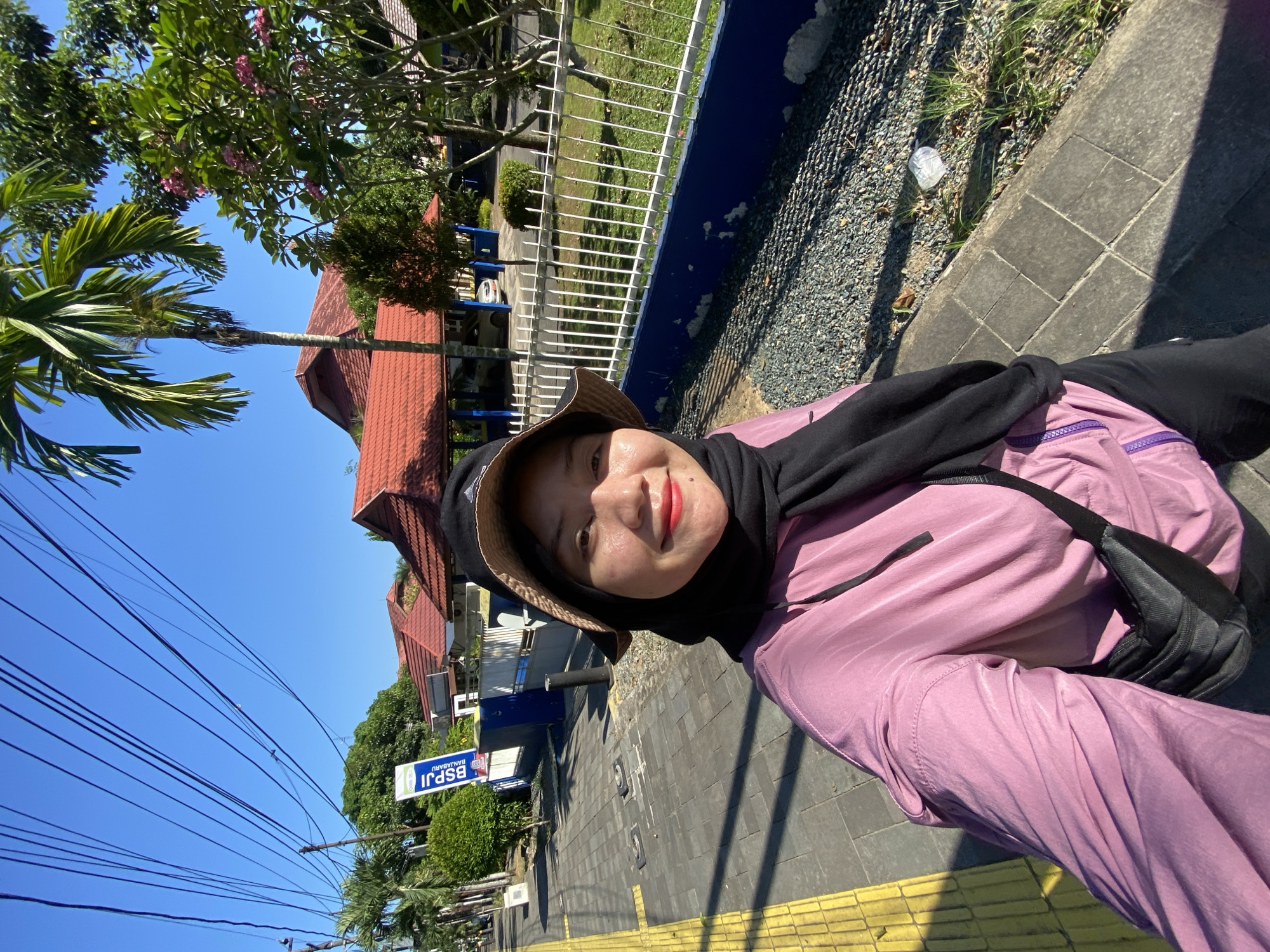 A woman in a pink hooded jacket, black hijab, and a bucket hat takes a selfie on a sunny day. She is wearing red lipstick. The woman is standing on a sidewalk with yellow tactile paving. Behind her is a white metal fence, a lawn, and a building with a red tiled roof. A sign that reads "BSPJ BANJARBARU" is visible on the left. Tall palm trees and other greenery are present in the background. The sky is clear and blue. The image appears to have been taken in the city of Banjarbaru, Indonesia.