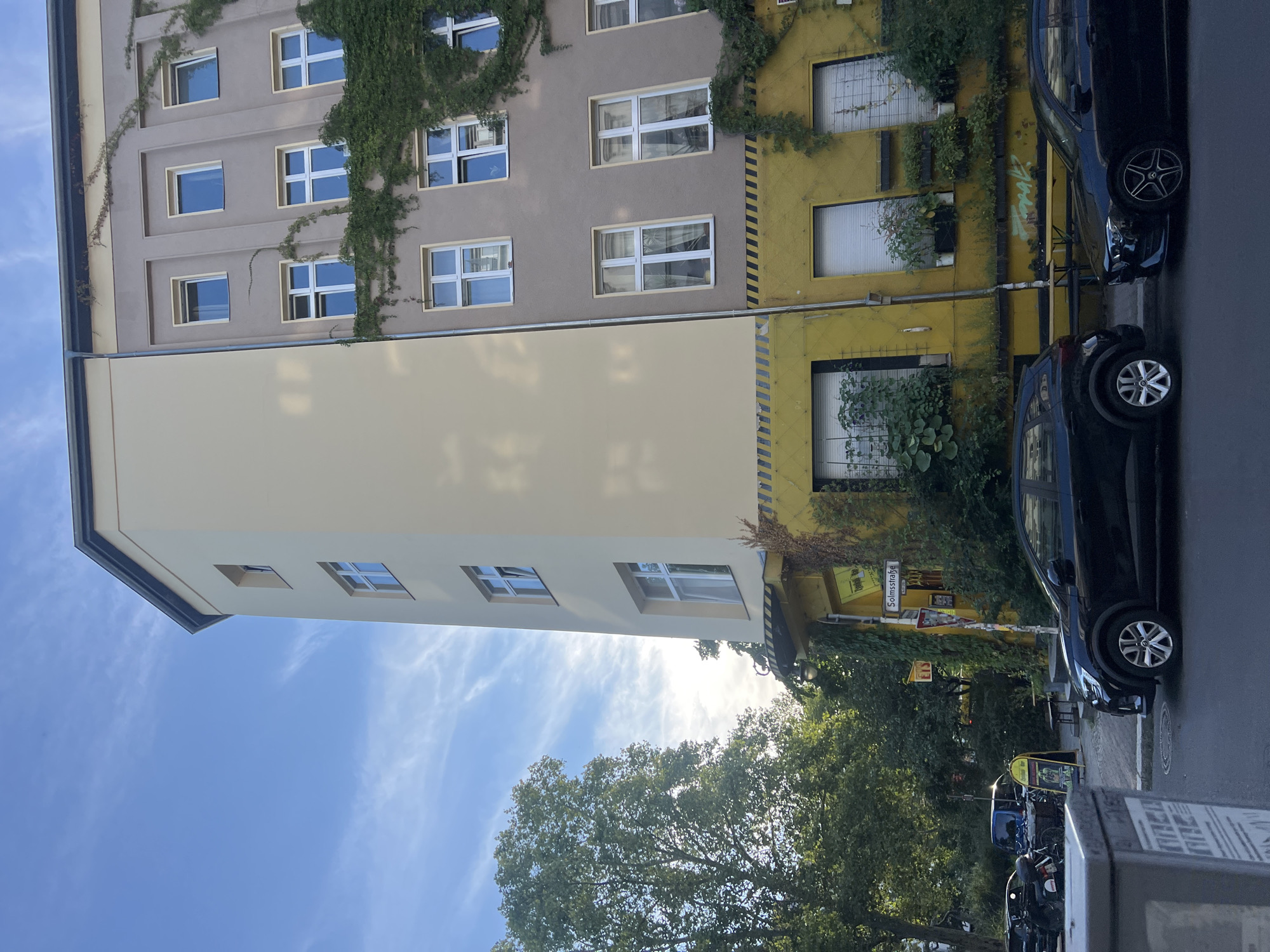 The image depicts an exterior view of a building with a distinct architectural style, located on Solmsstraße in Berlin, Germany. The building exhibits a curved facade on the left, painted a pale yellow, and a more angular section on the right, painted a muted brown with yellow accents on the lower floors. Green ivy climbs up the right side of the building, partially obscuring the windows. Several windows, predominantly white-framed and multi-paned, are visible on both sections of the building.
At street level, the yellow section of the building features large windows with white blinds and planters filled with greenery. A street sign for "Solmsstraße" is mounted on a pole. In front of the building, a black SUV is parked on the street, facing towards the right. To its right, another black car, a sedan, is also parked. The street appears to be paved with asphalt, and a sidewalk is visible. Trees and other foliage line the street, suggesting a well-treed urban environment. The sky above is blue with wispy white clouds, indicating daylight. The overall scene conveys a sense of urban life in a European city.