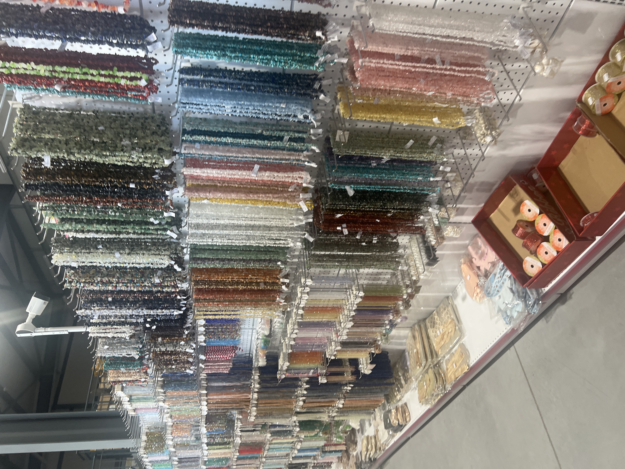 The image shows the interior of a store, likely a craft or bead shop. Numerous strands of beads in various colors and materials hang from display racks. The beads are organized by color, including shades of red, green, blue, yellow, brown, and white. Other craft items, possibly ribbons and embellishments, are displayed on shelves near the beads. There are no people visible in the frame. The store is located in Burgas, Bulgaria.