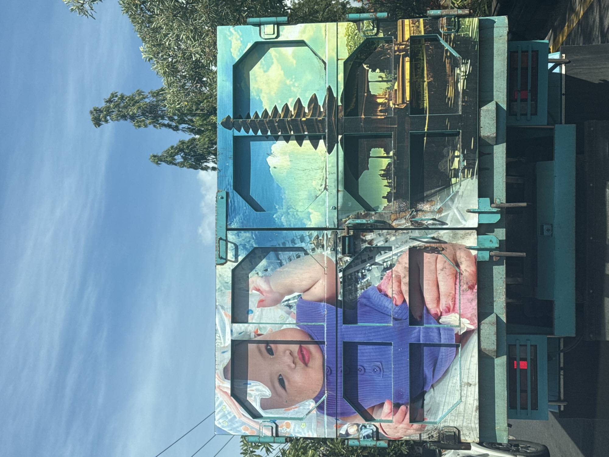 A teal truck in Denpasar, Indonesia, is adorned with images spanning its container doors. The top half of the doors displays a pagodalike structure set against a cloudy sky on the left, mirrored with a tan-colored cityscape on the right. The lower half of the doors features a baby with a blue shirt and pink blanket with hands, presumably of an adult, holding a wipe. The sky is visible behind the truck with a tree in the upper left. A road can be seen on the right behind the truck, and the back of a car appears at the bottom of the frame.