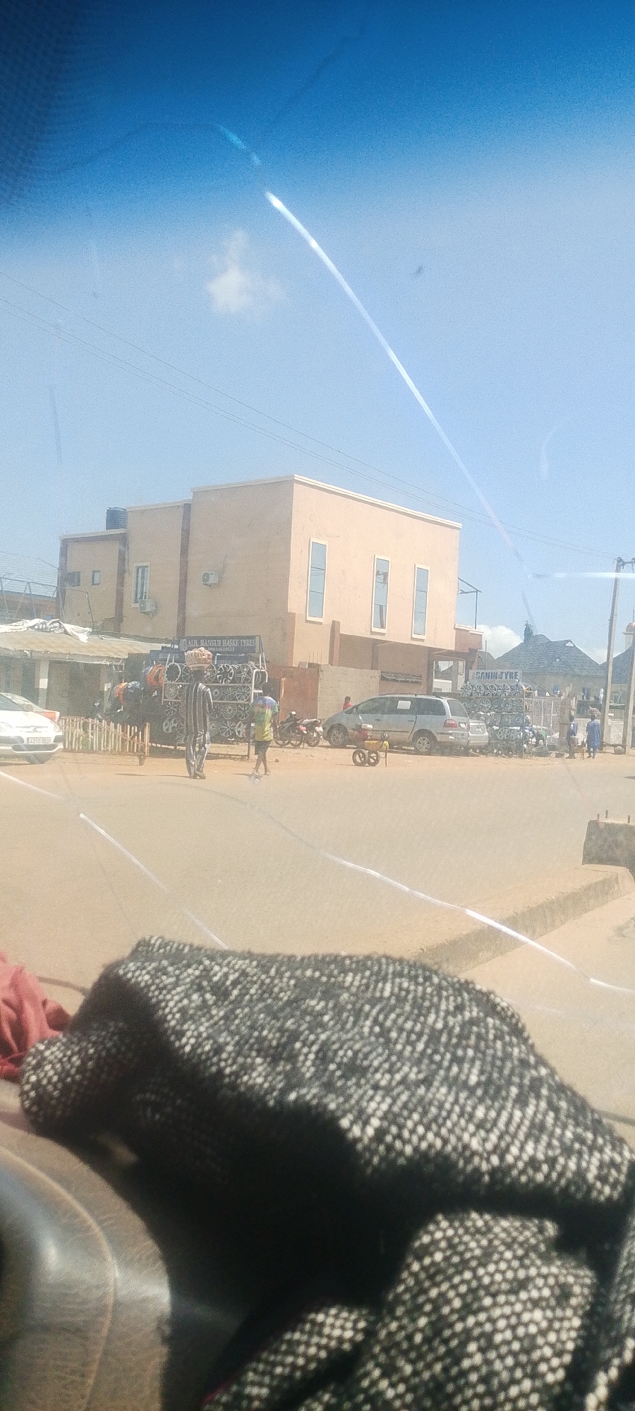 The image depicts a street scene in Jos, Nigeria, on a clear day with a bright blue sky. The foreground is dominated by a dark, patterned fabric, possibly a blanket or clothing, resting on a car's interior.
In the mid-ground, a two-story building with a light beige facade is visible. The building has several windows with dark frames and what appear to be air conditioning units mounted on the walls. In front of this building, there is a stall or shop selling tires. The tires are stacked in large quantities, forming a visually prominent display. A sign above the tire display reads "ALI HASSAN HASKE TYRES". Next to this, another sign with similar tire displays shows "SANIN TYRE".
Several people are present in the scene. A person wearing a striped shirt is standing near the tire display, possibly a vendor. Another person in a green and yellow shirt is seen with a broom, appearing to sweep the ground. A few motorcycles are parked near the tire stall. A silver minivan is parked on the road, and another white car is visible on the left side of the image.
In the background, more buildings and structures are scattered across the landscape, suggesting a built-up urban or semi-urban area. The overall impression is of a busy commercial street. Light flares and reflections from the camera or windshield are visible across the image, particularly in the sky and on the road surface.