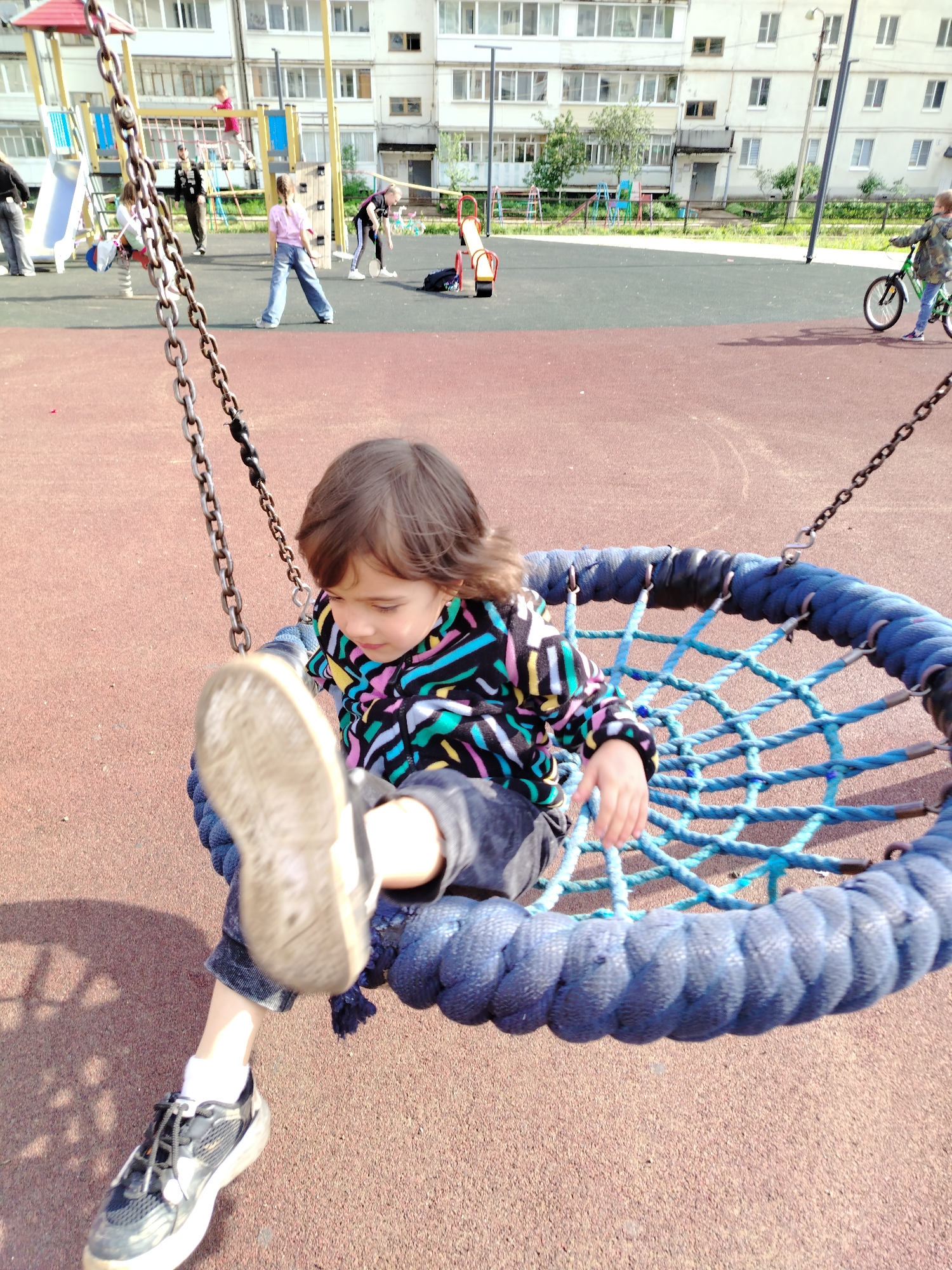 A young child sits in a blue rope swing in a playground. The child has brown hair and is wearing a colorful, patterned jacket, dark jeans, and mismatched shoes - one with a visible sole and the other a sneaker. The playground surface is red. In the background, other children are playing on various playground equipment including slides, a seesaw, and a climbing structure. A person on a bicycle is visible to the right. A multi-story building stands behind the playground, which suggests an urban environment. Based on prior knowledge, this scene is located in Noviy, Russia.
