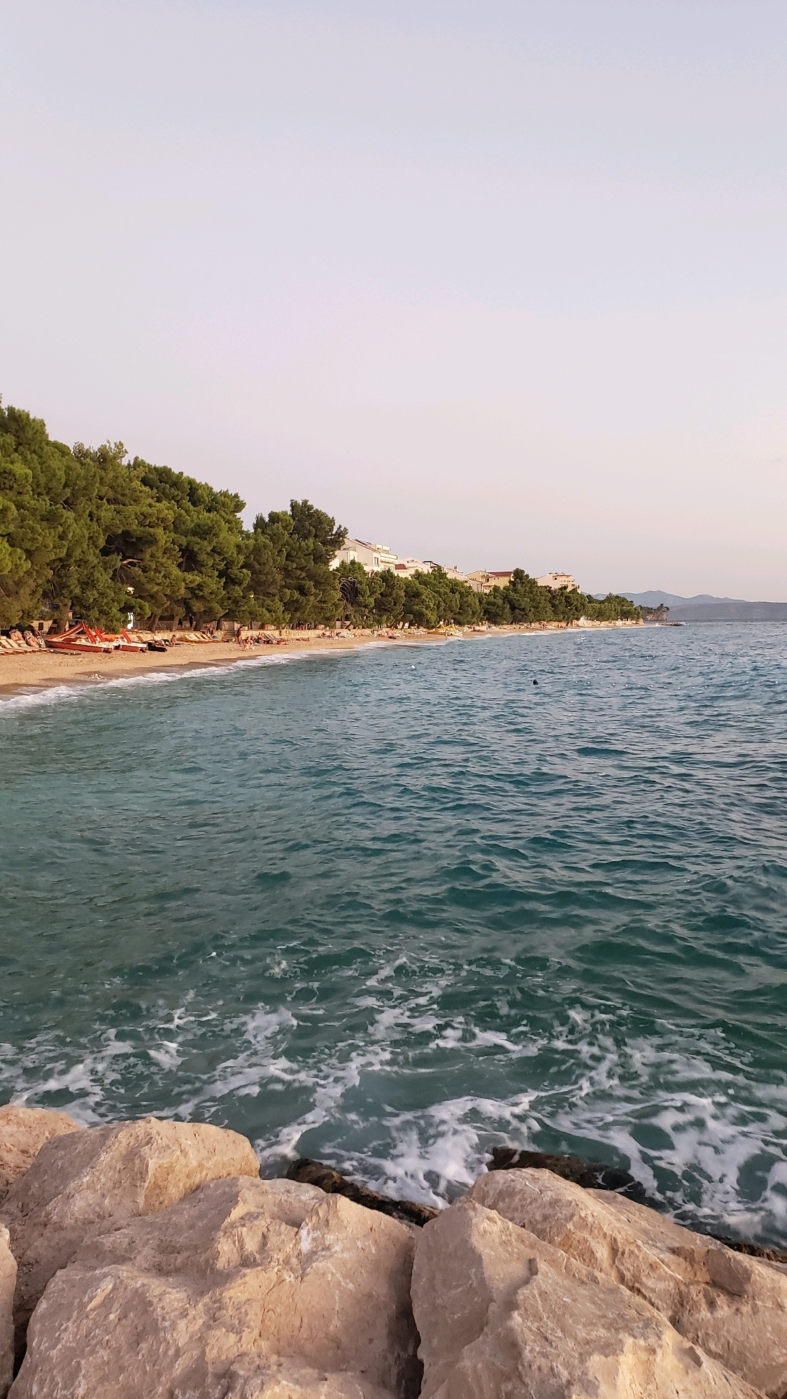 The image depicts a coastal scene in Tučepi, Croatia, characterized by a sandy beach lined with pine trees and a few buildings. The sea is a vibrant turquoise with gentle waves lapping against the shore and foam churning around a rocky embankment in the foreground. Beach chairs and umbrellas are scattered along the sand, with a few sunbathers visible. Red rescue boats are also present on the beach. The sky is a soft, pale hue, suggesting it is either early morning or late afternoon. In the background, distant hills are faintly visible on the horizon. The overall impression is one of a serene and pleasant beach environment.