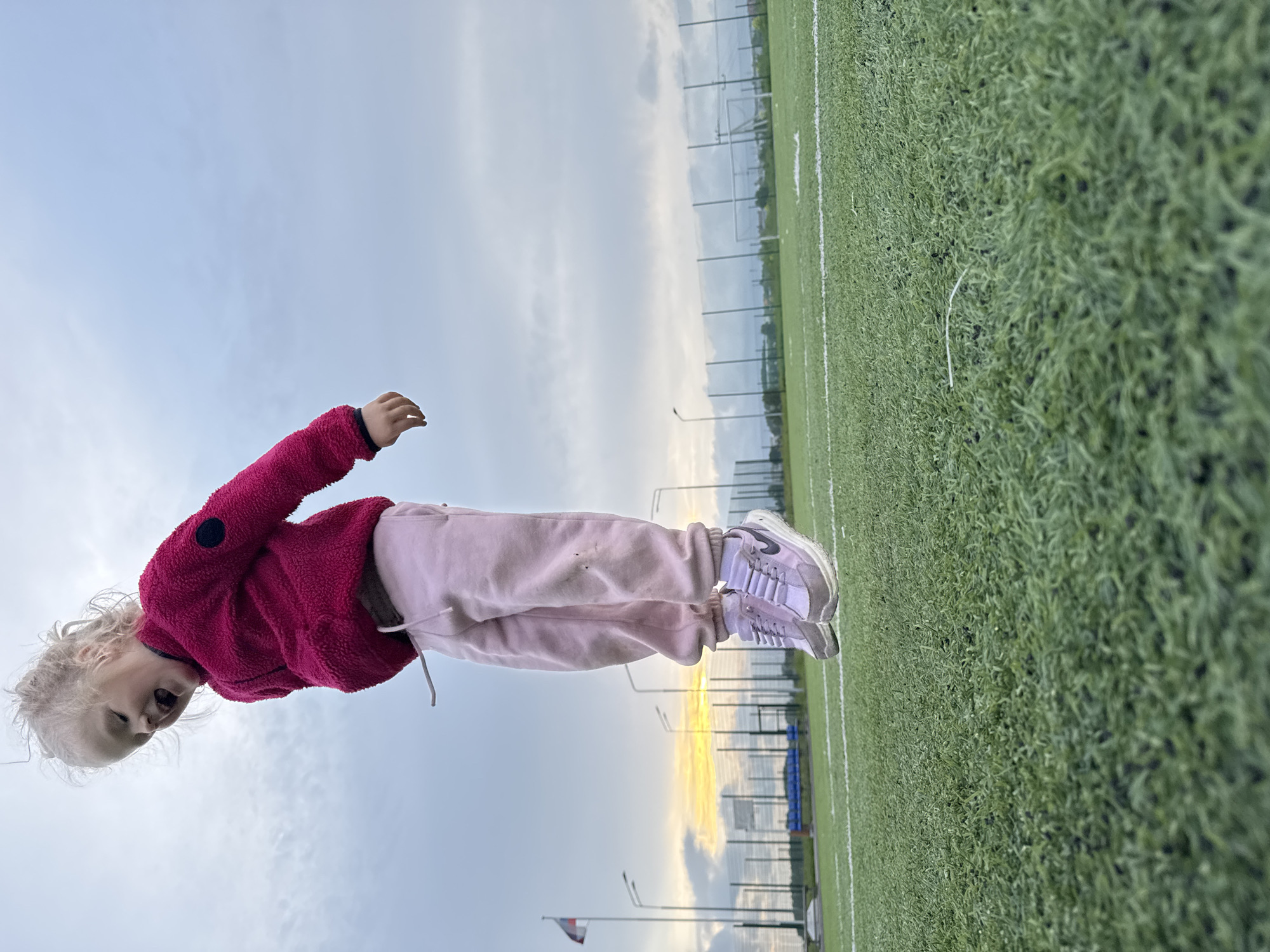 A young blonde child is captured mid-air, suspended a few inches above an artificial green turf sports field with white markings. The child, wearing a bright pink fleece jacket with a dark circular patch on the left arm, light pink sweatpants with a visible white drawstring, and light purple Nike sneakers, has an open mouth and appears to be laughing or shouting, looking down and to the left. The scene is located in the city of Ферма, Russia. The background shows an overcast sky with patches of grey-blue clouds, transitioning to a warm orange and yellow sunset glow on the horizon to the left. Visible environmental elements include a flagpole flying a Russian flag on the far left, multiple tall streetlights, perimeter fencing, blue benches beyond the fence, and a white soccer goalpost in the distance on the right.