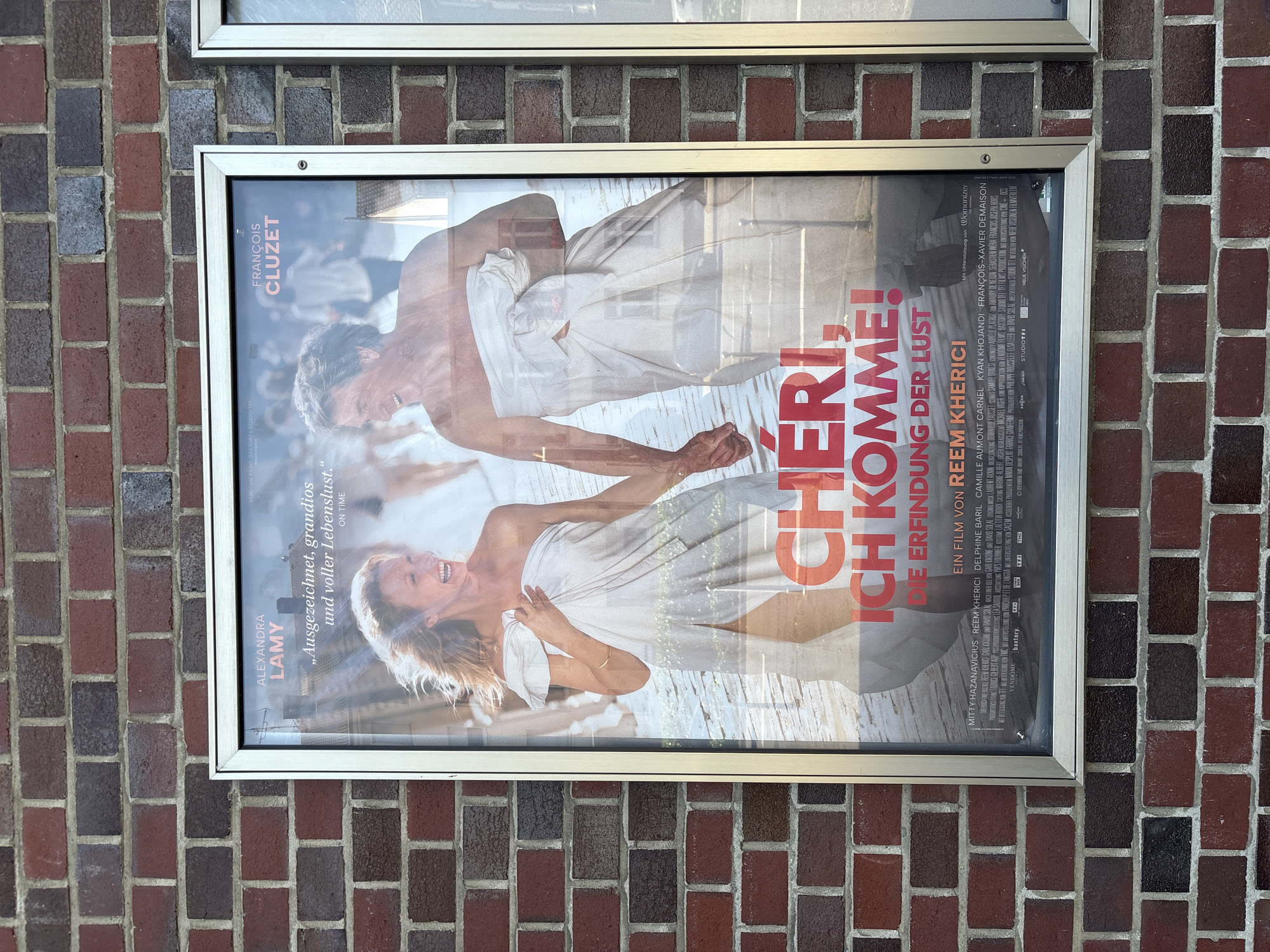 A movie poster is mounted on a red brick wall. The poster advertises the film "Chéri, ich komm'e! Die Erfindung der Lust" (Cheri, I'm coming! The invention of lust). The poster features a close-up shot of two adults, a man and a woman, holding hands and wrapped in white sheets. They are smiling. The title of the film is written in large, red, sans-serif font. The names of the lead actors, Alexandra Lamy and François Cluzet, are printed above their respective images. The poster also includes a director's credit for Reem Kherici. The movie poster is enclosed in a silver frame. The wall is constructed of weathered red bricks with dark mortar. The image was taken on June 15, 2024, in Mönchengladbach, Germany.