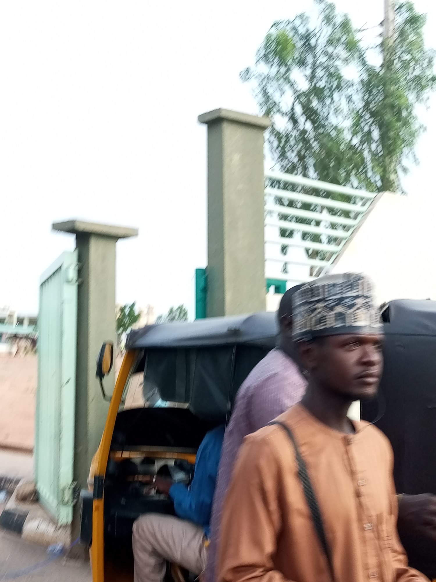 This is a medium-distance, eye-level shot of a street scene in Gombe, Nigeria, likely taken near a gateway. In the foreground, a person wearing an orange, buttoned tunic and a patterned Kufi cap is partially visible on the right, facing away from the camera and looking to the right. A portion of another person in a pink shirt is visible to the left of the person in orange. Behind them, the driver of a yellow auto-rickshaw, wearing a blue shirt and light-colored trousers, is seated and appears to be operating the vehicle. The auto-rickshaw's interior is dark, with a steering wheel visible. The background features a light-colored, open gateway with a green gate on the left. Further back, a white railing or fence is visible, and a tree with green foliage is present in the upper right quadrant of the image. The sky is overcast and white. The scene suggests a moment of transit or waiting within the urban environment of Gombe.
