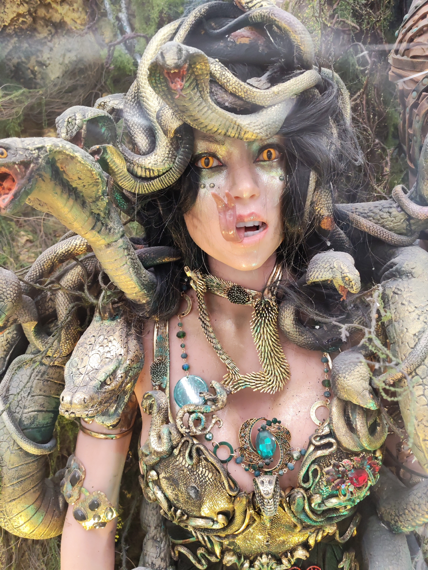 This is a medium shot, eye-level, full-body shot of a Medusa costume. The costume features a headpiece composed of numerous snakes, some with their mouths open and tongues out, giving a menacing appearance. The subject has striking yellow eyes and her mouth is open, with a partially visible tongue. She is adorned with intricate, golden armor and elaborate necklaces featuring gemstones in blue and red. Snakes are also integrated into the armor and surrounding her body. The background is blurred and appears to be a natural, outdoor setting with foliage. The lighting is somewhat dramatic, with highlights and shadows emphasizing the details of the costume and the subject's features.