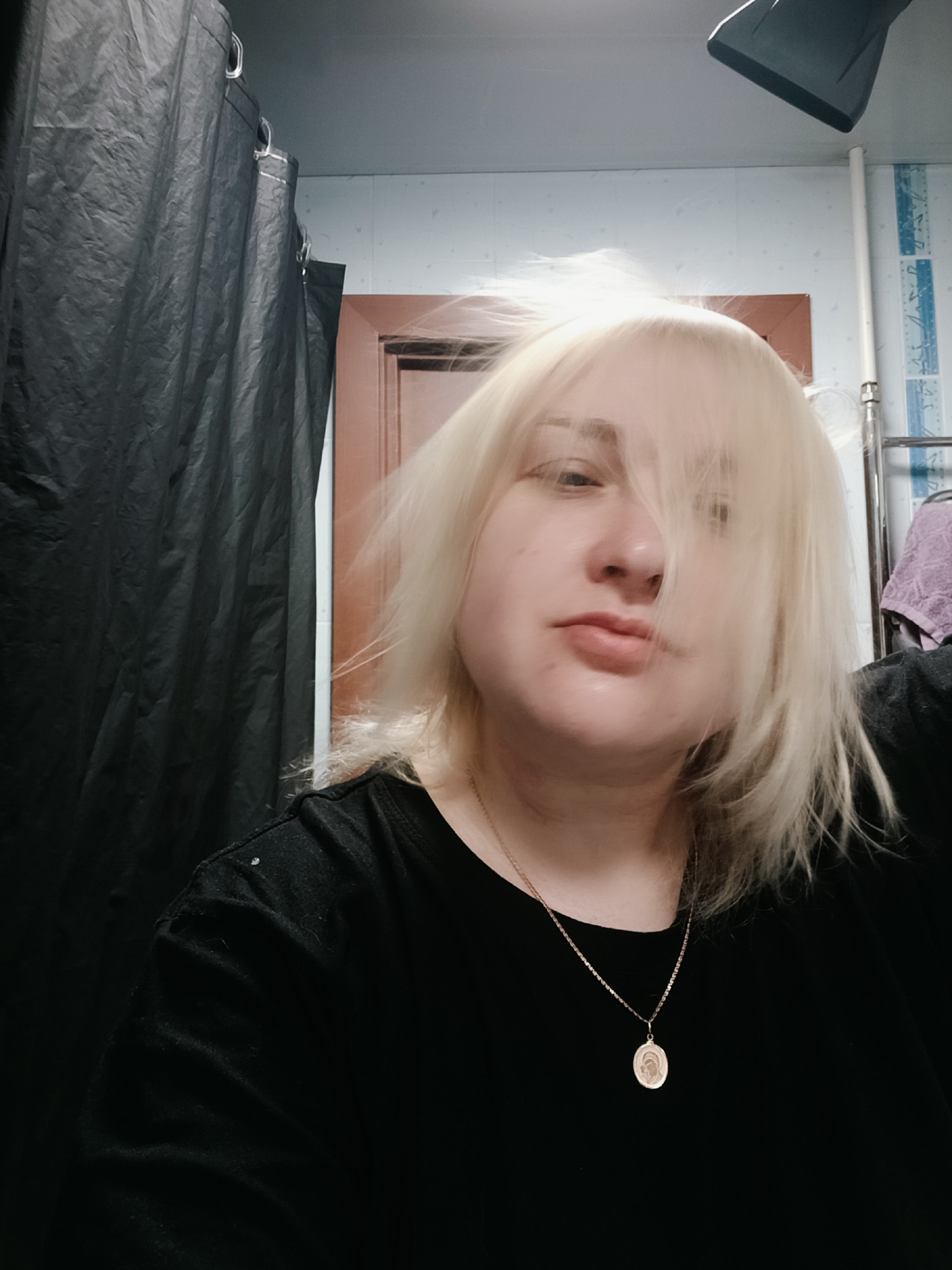 The image appears to be a selfie taken in a bathroom setting. A woman with fair skin and bleached blonde, somewhat disheveled hair is in the foreground, wearing a black t-shirt and a gold necklace with a pendant. Her expression is neutral. Behind her to the left is a dark, possibly black, shower curtain. In the background, elements of a bathroom can be seen, including white-blue patterned tiles and a wooden-framed door. There also appears to be a light fixture above her head. The location is presumed to be in the city of Chaikovsky, Russia, based on the prompt's instructions, but there are no visual cues to confirm this definitively within the image.