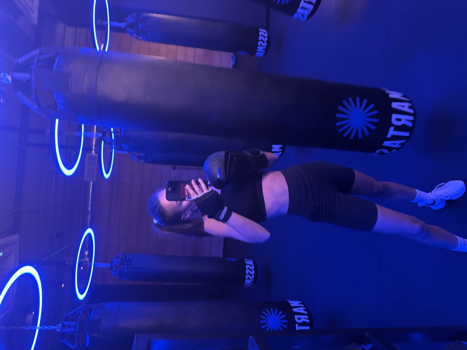 A woman takes a mirror selfie in a boxing gym. She's wearing boxing gloves and wrist wraps, a black sports bra, and black biker shorts, plus white sneakers and socks. Behind her are several black punching bags hanging from the ceiling, illuminated by blue lighting and neon ring lights. The gym floor is also blue. The image is slightly tilted to the right.